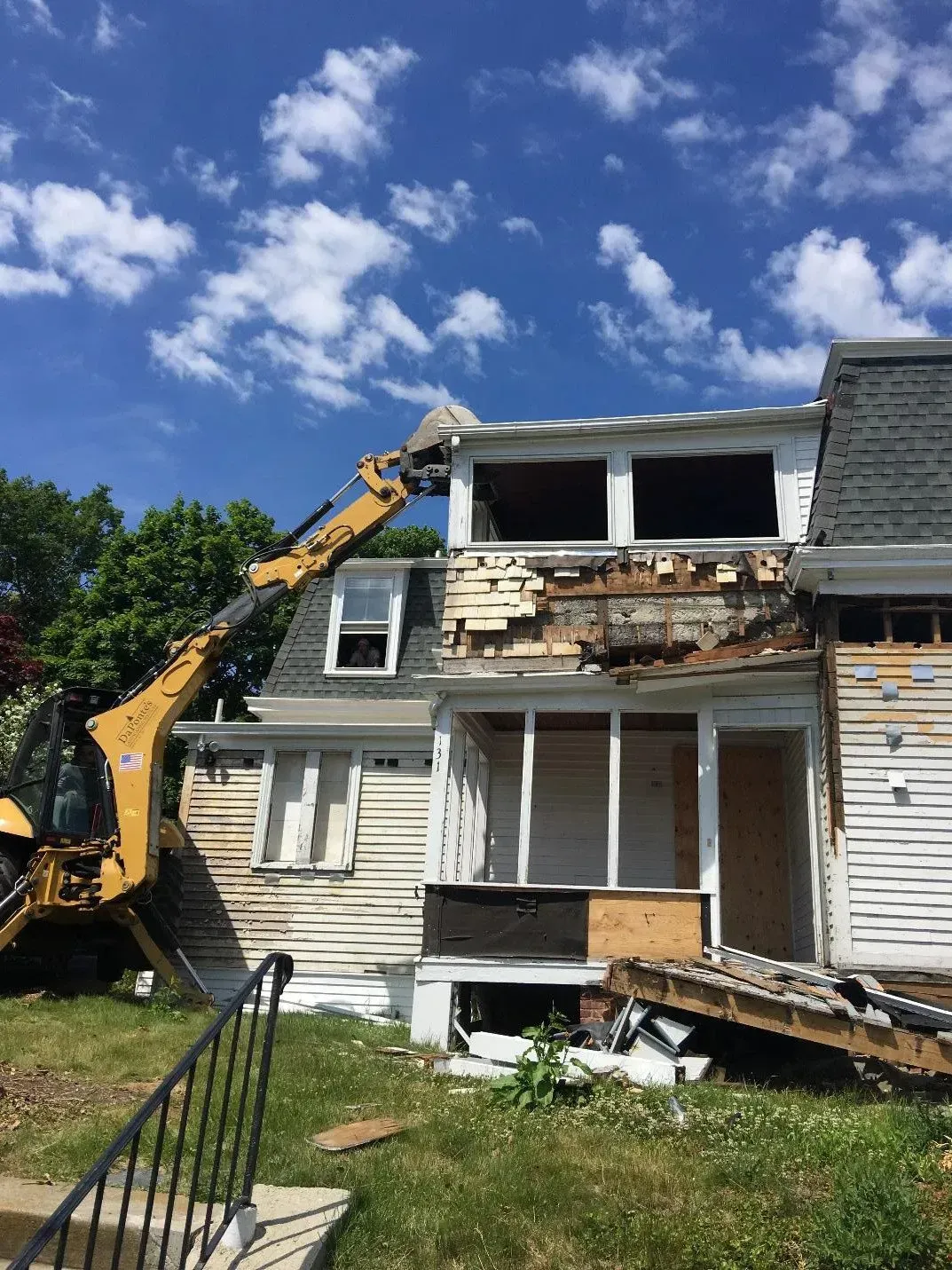 A yellow excavator demolishing a weathered, two-story house under a blue sky.