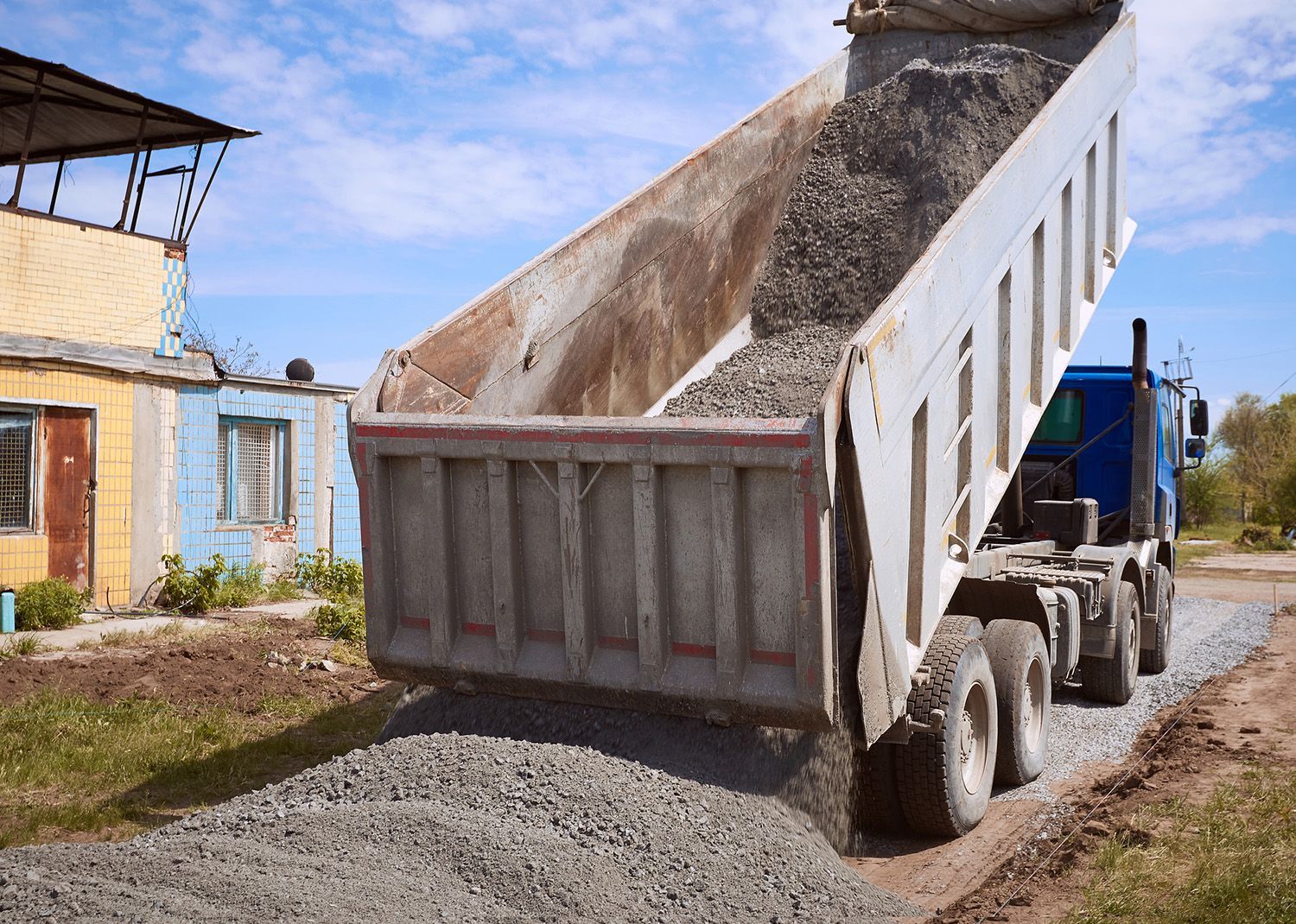 Dump truck unloading gravel onto a road near a small building on a sunny day. Dump truck unloading gravel onto a road near a small building on a sunny day.