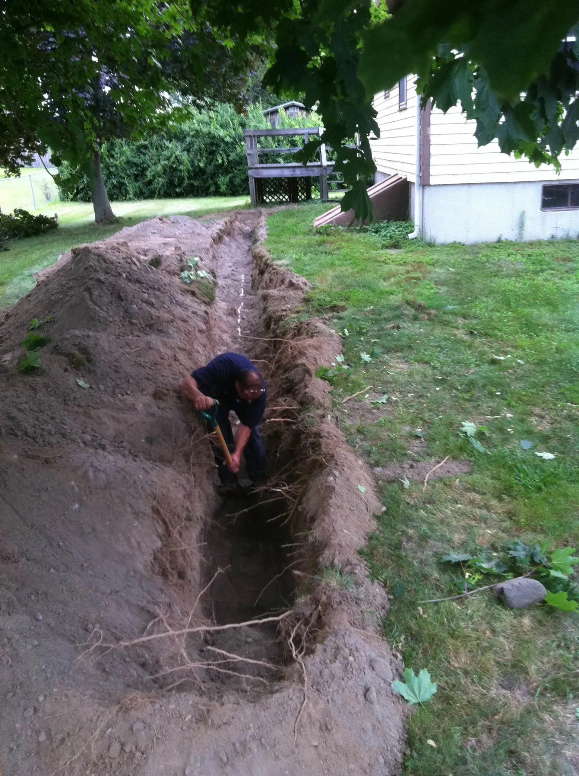 Person digging in a trench in a grassy yard, near a house and trees. Person digging in a trench in a grassy yard, near a house and trees.