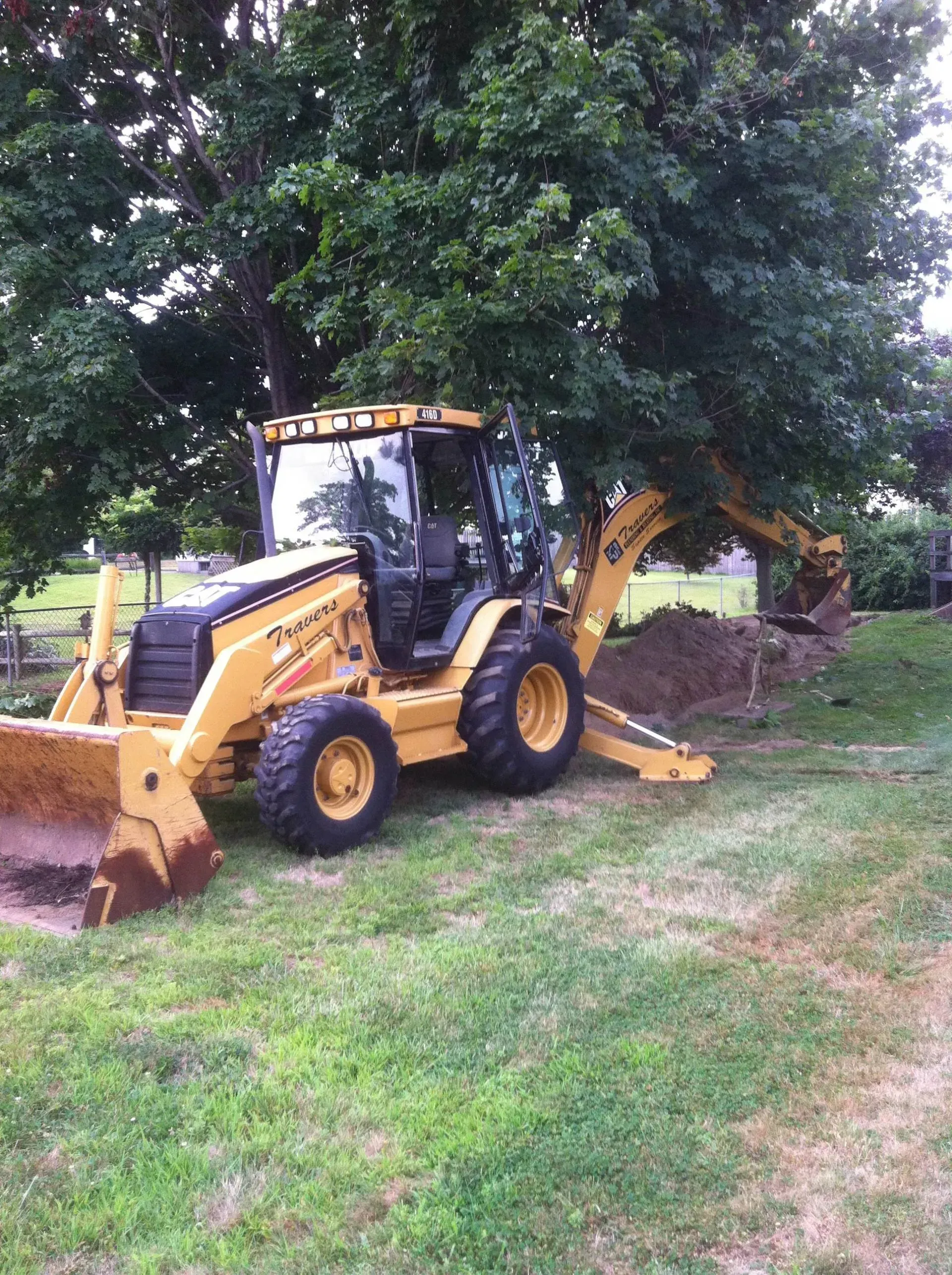 Yellow backhoe digging near a tree in a grassy yard.