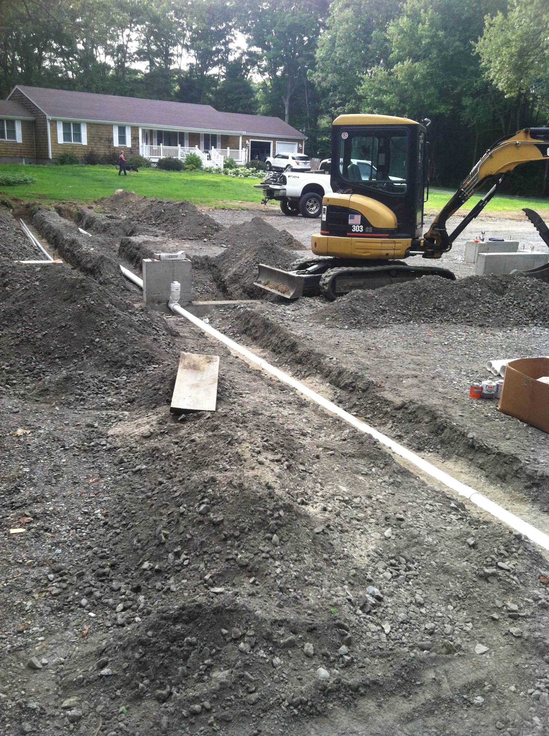 Construction site with mini excavator digging trenches, laying pipes in front of a residential house.