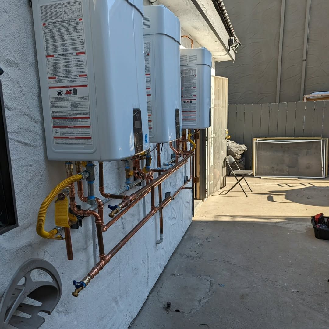 Three white water heaters mounted on an outdoor wall, connected by copper pipes and gas lines.