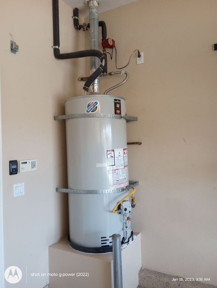 Tall, cylindrical water heater in a utility room. Gray tank with attached pipes and wiring, set against a beige wall.