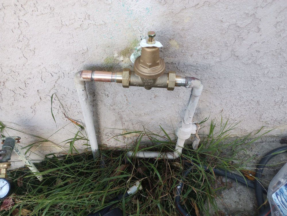 Water valve and pipes against a textured wall, surrounded by overgrown grass.