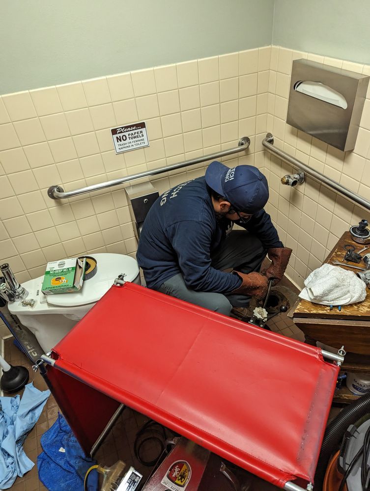A person in a blue shirt and hat working on plumbing in a tiled bathroom, with a red cart in front.