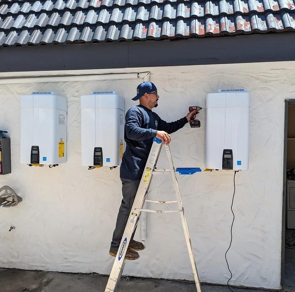 A person on a ladder installs white water heaters on an exterior wall.