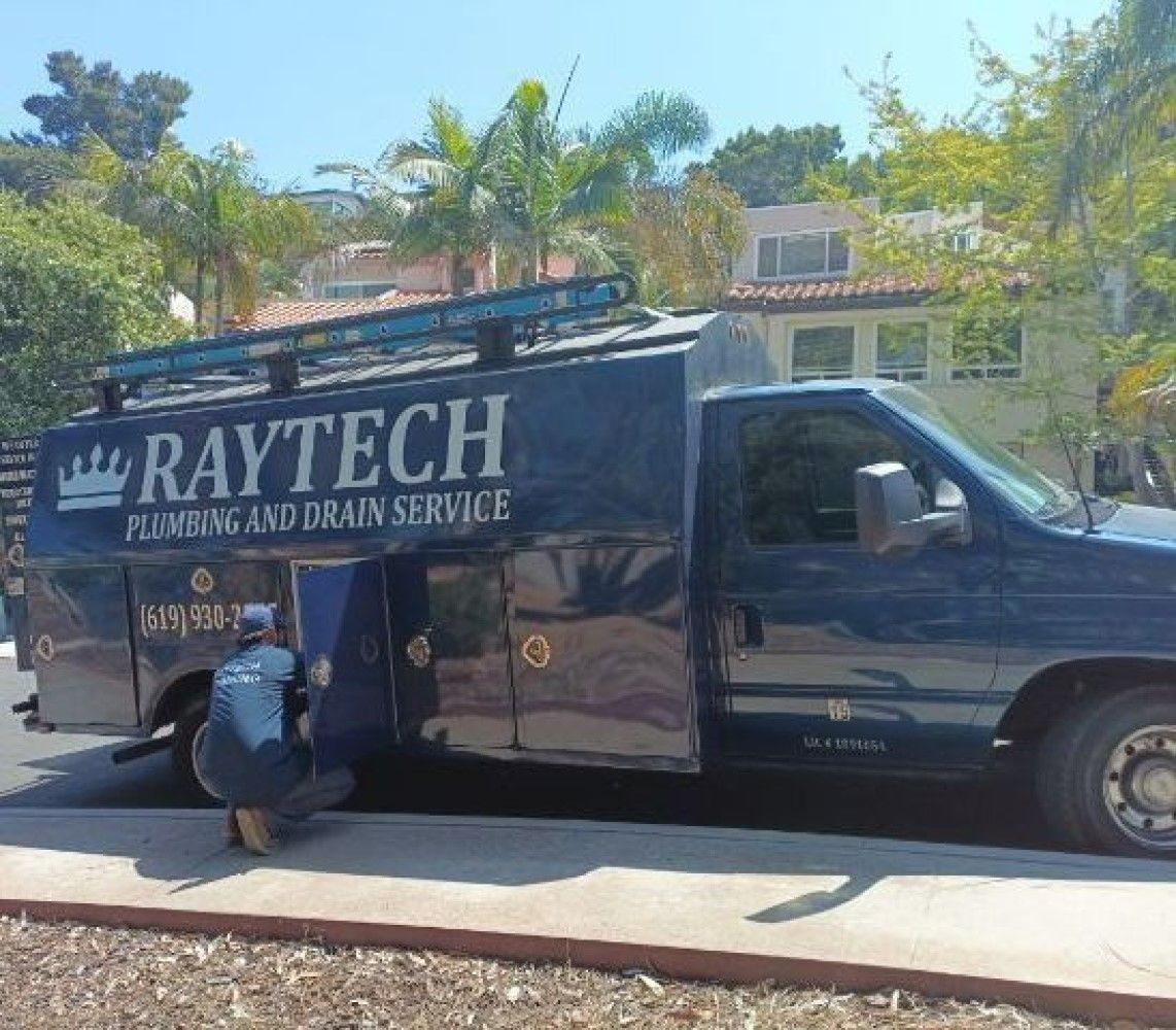 Raytech plumbing service truck with a worker kneeling, opening a side compartment, in a residential area.