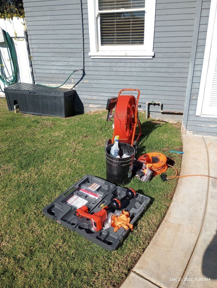 Plumbing equipment set up on green lawn near a gray house: a reel, tools, and bucket.
