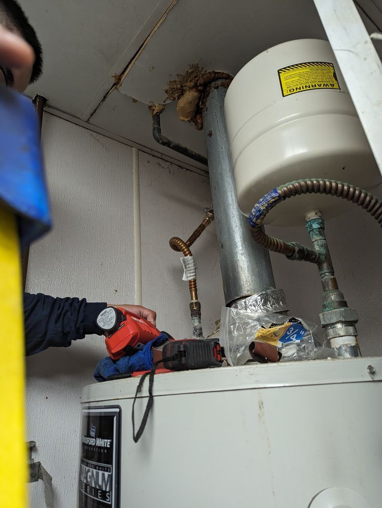 Person examining a water heater in a utility room; pipes, tank, and tools are visible.