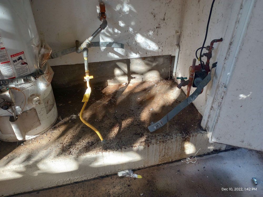 Water heater and pipes in a small concrete room with debris on the floor and a yellow electrical cable.