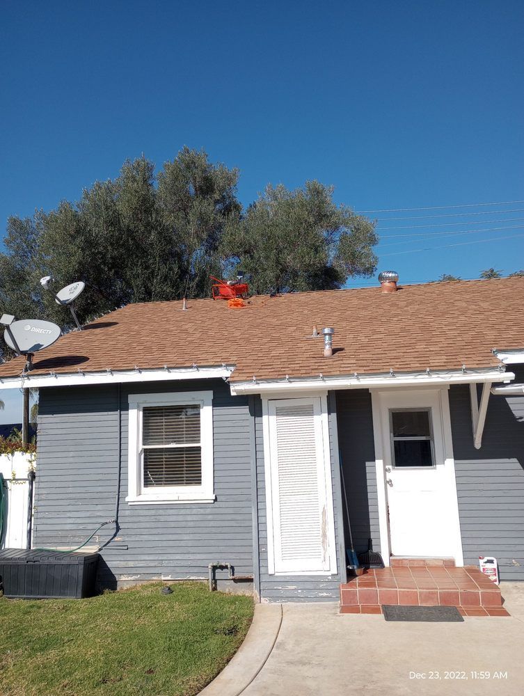 Small blue house with brown roof and white trim. Satellite dishes and trees are visible. Clear, sunny sky.