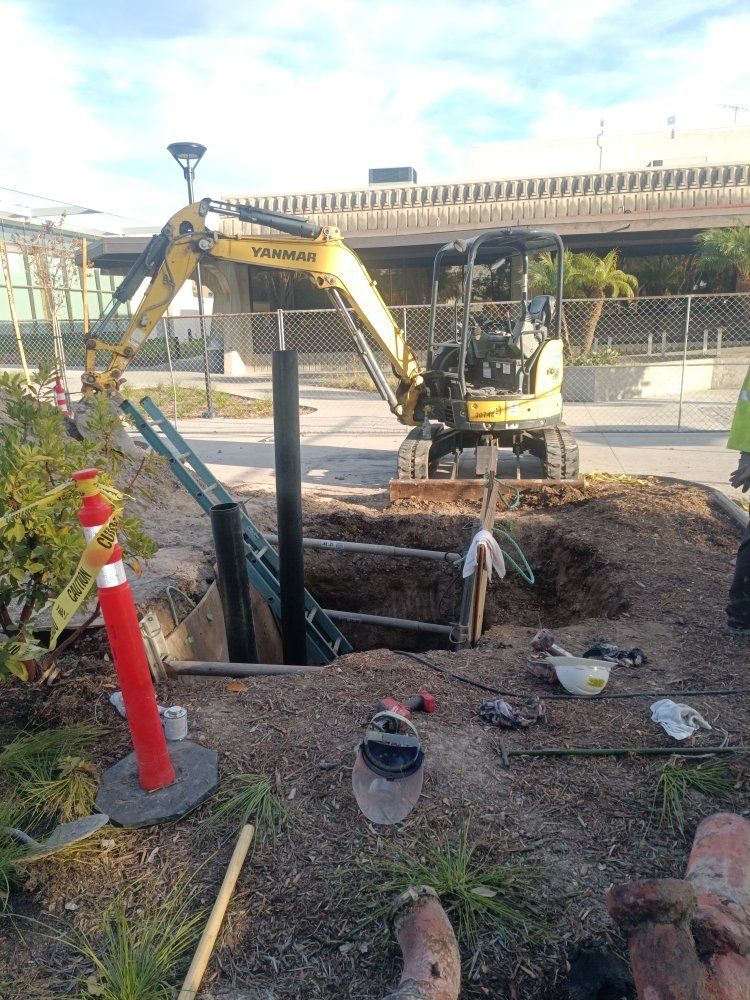 A small yellow excavator digs in a dirt pit. Black pipes and tools are nearby; a red post is in the foreground.