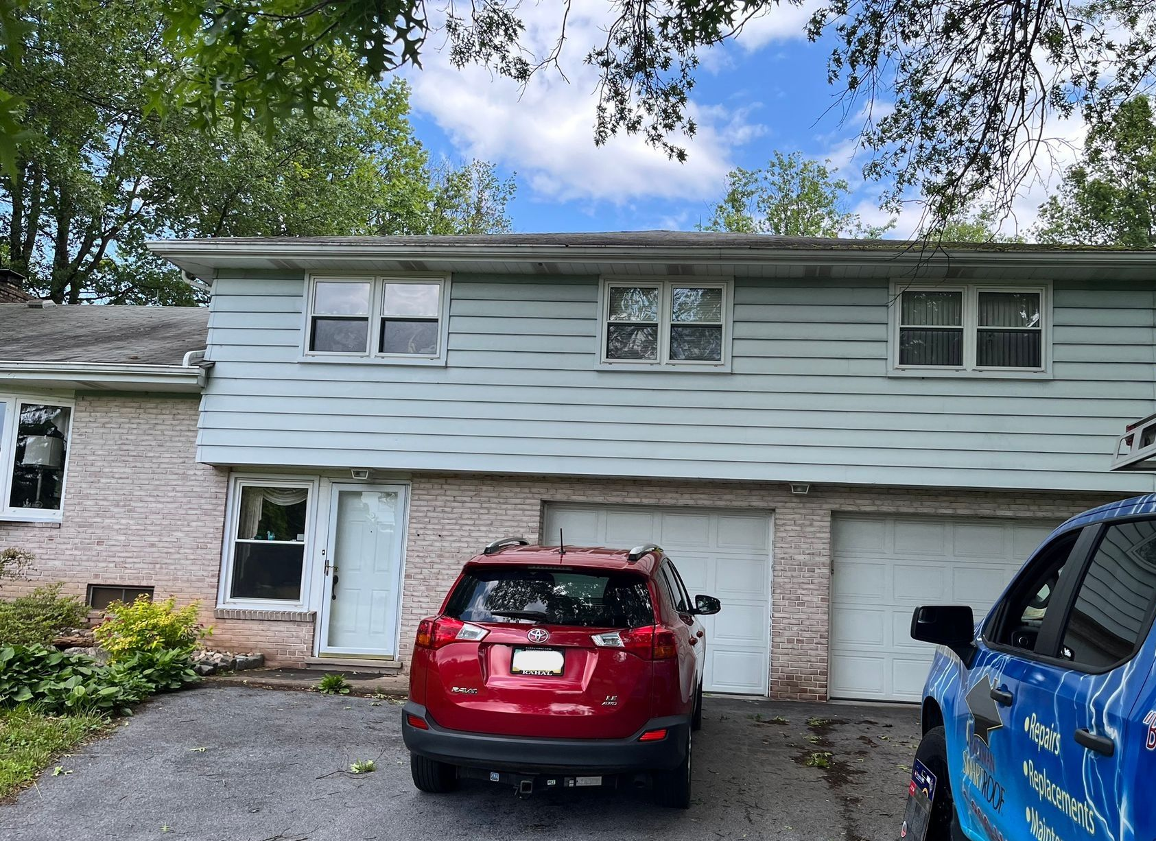 A two-story house with light blue siding and a light stone lower level, featuring a red SUV parked in the driveway.
