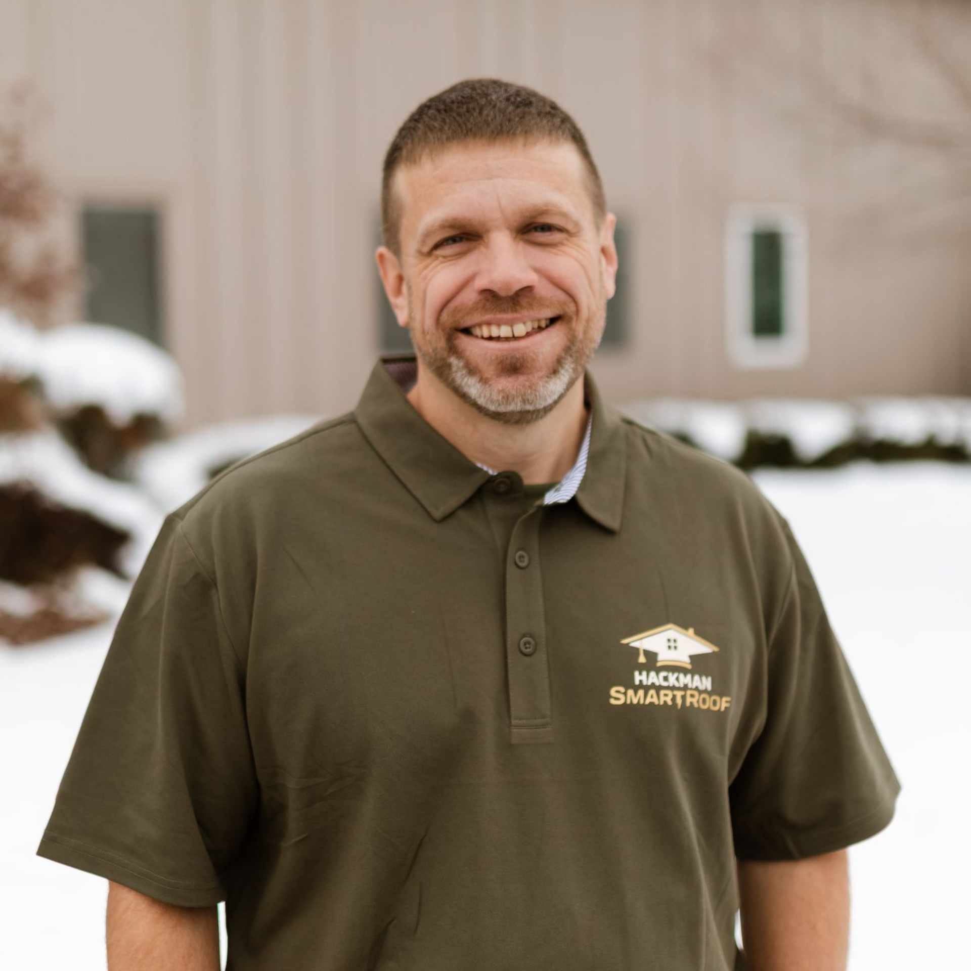 A smiling man in a dark green branded polo shirt stands outdoors in a snowy setting.