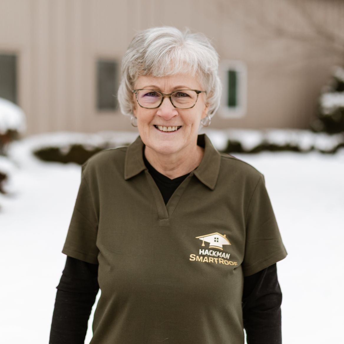 A smiling person with gray hair and glasses wearing an olive green polo shirt outdoors with snow in the background.