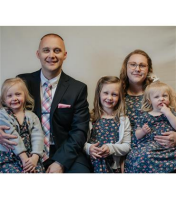 Family of five smiles at the camera: man in suit, woman in floral dress, and three young girls.