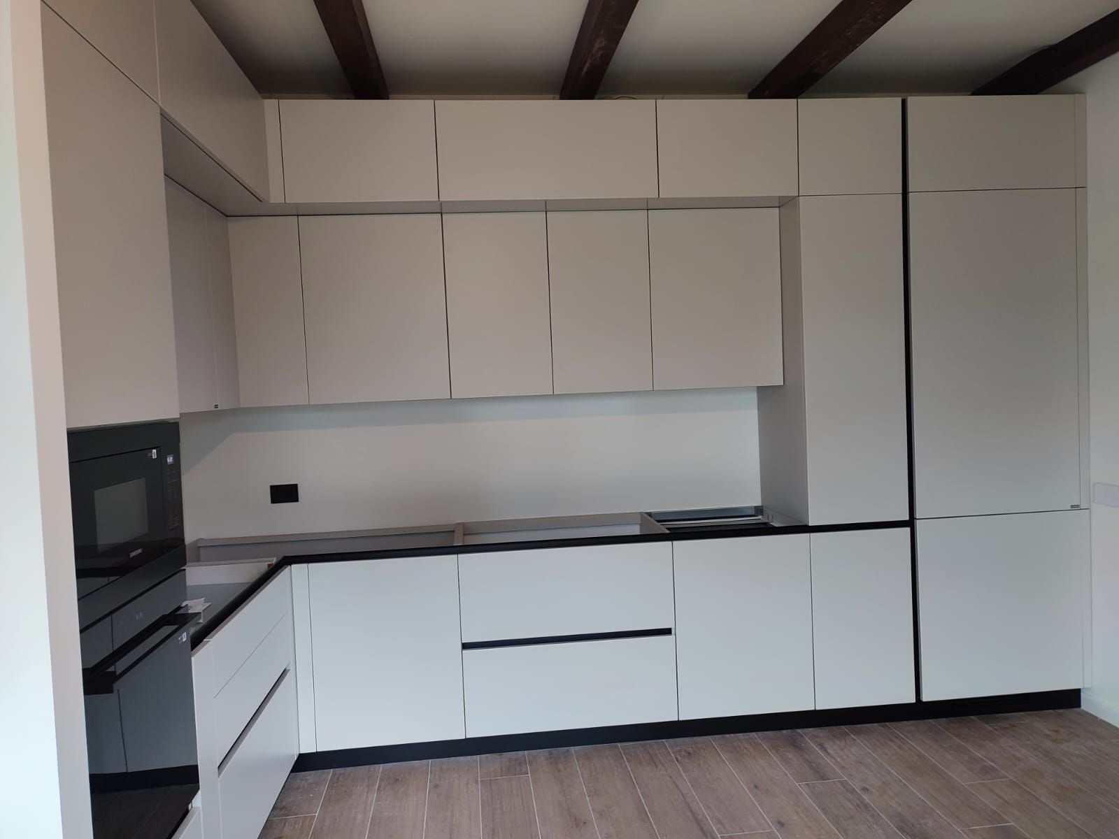 Moderna cucina bianca con dettagli neri, dotata di mobili, elettrodomestici e travi in ​​legno sul soffitto.