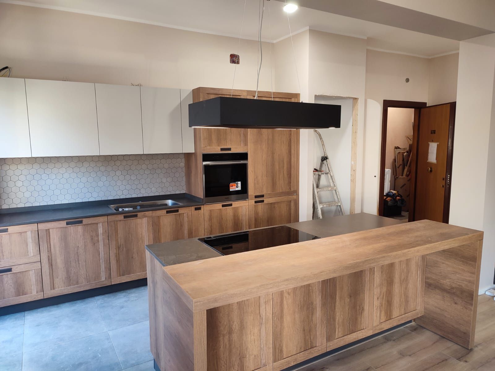 Cucina moderna con mobili in legno, isola e cappa nera; è visibile una porta.