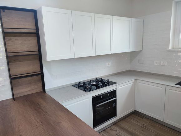 Cucina bianca con mobili, piano cottura e forno. Ripiani in legno sul lato sinistro.