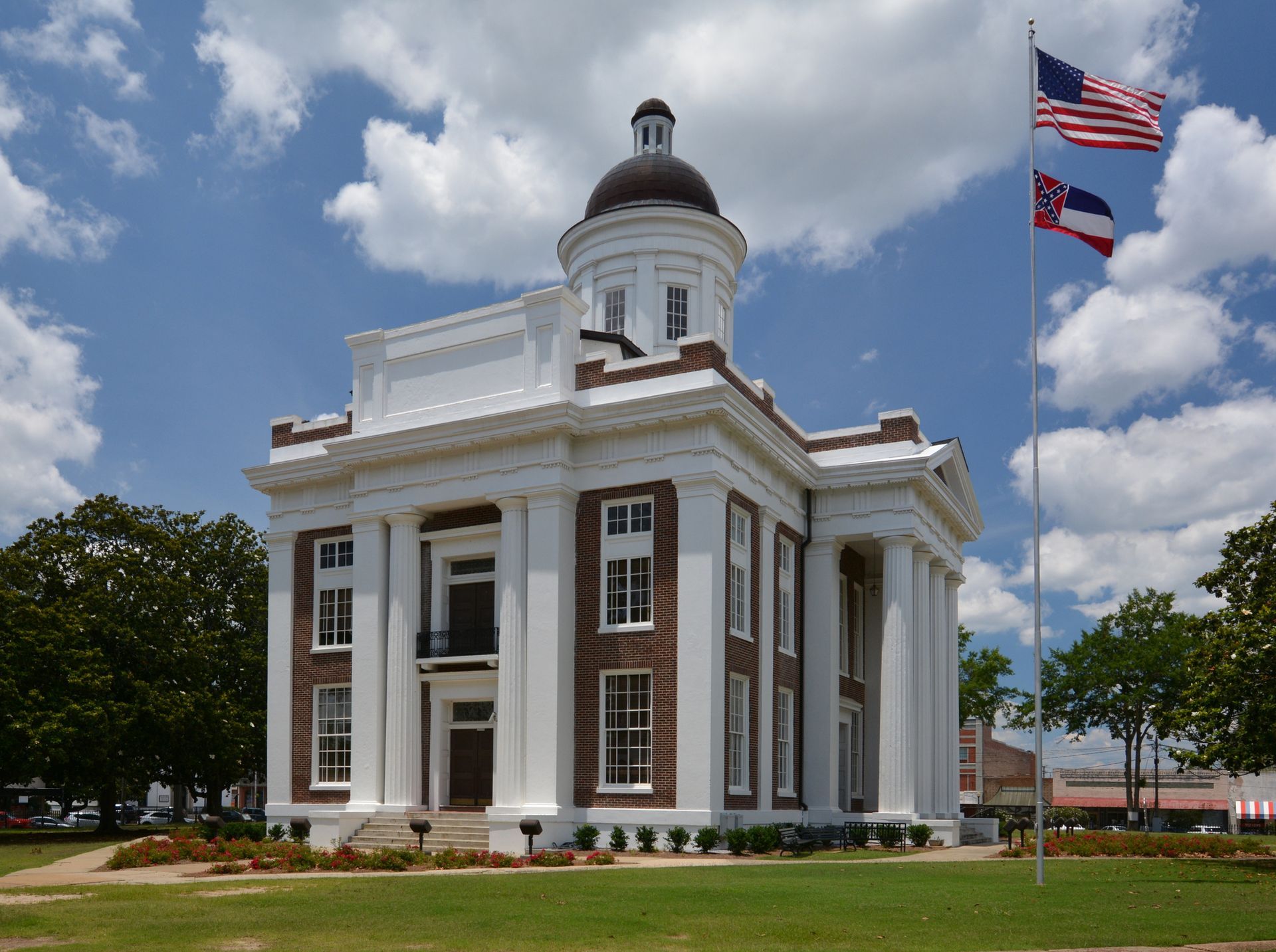 White courthouse with pillars, brick accents, and a dome under a cloudy sky. Flags fly.
