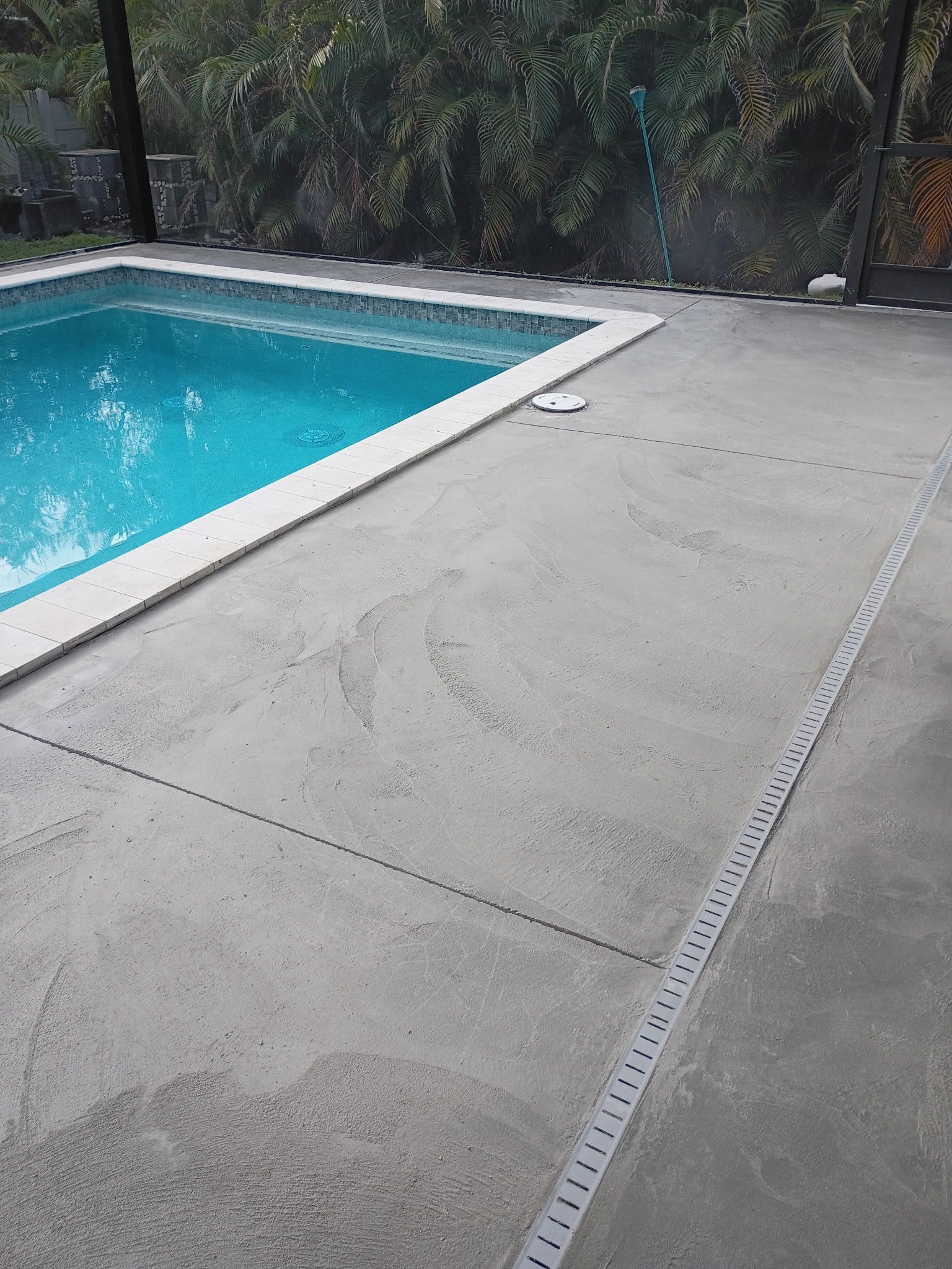 Concrete pool deck beside a turquoise swimming pool, with trees and a fence in the background.
