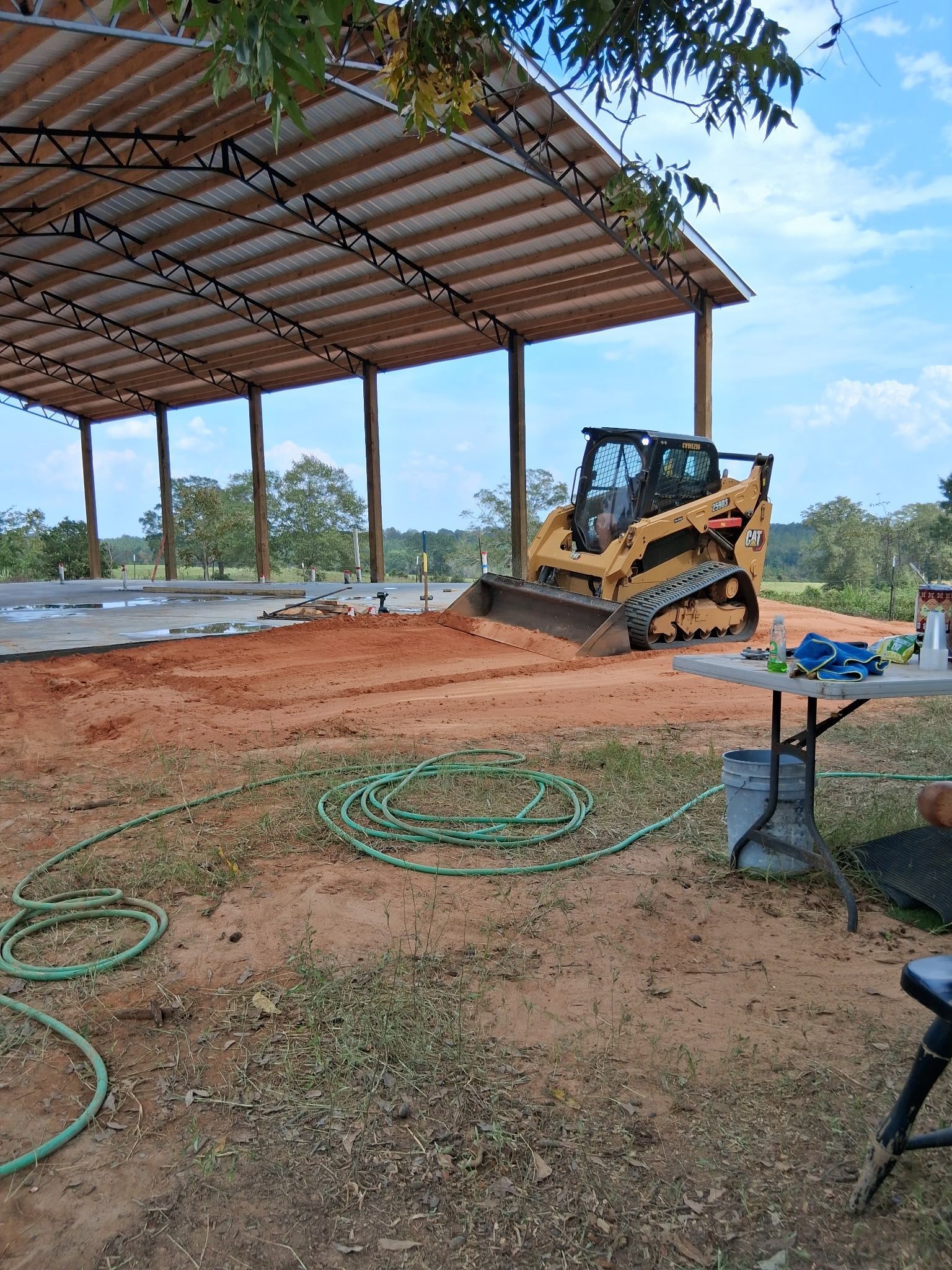 Skid steer grading red dirt under a metal-roofed structure. Green hose and a folding table with tools are visible.