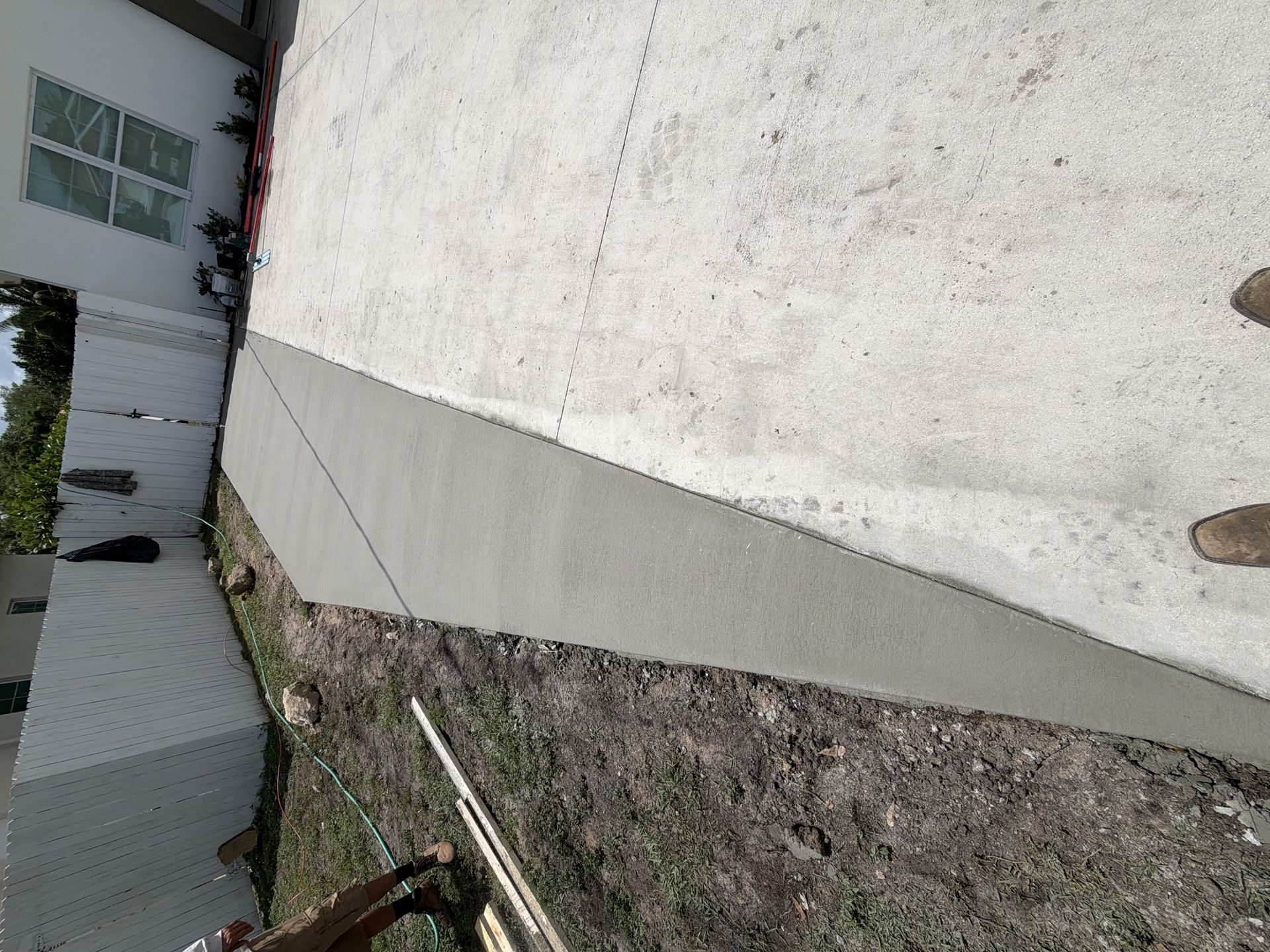 Concrete walkway beside a white building and fence, with dirt and grass along the edge.