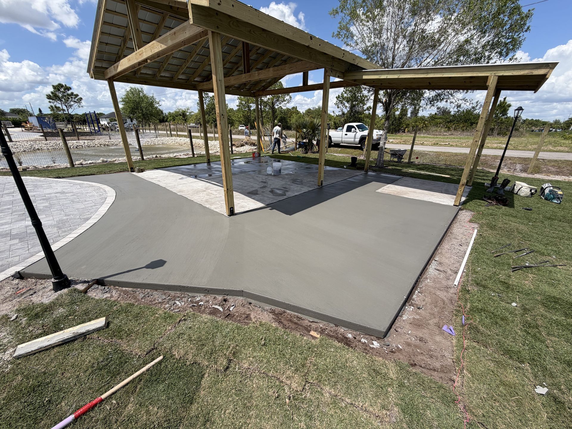 Concrete pavilion with wooden posts and metal roof on a grassy lot, beside a curved paved area and parked vehicles.