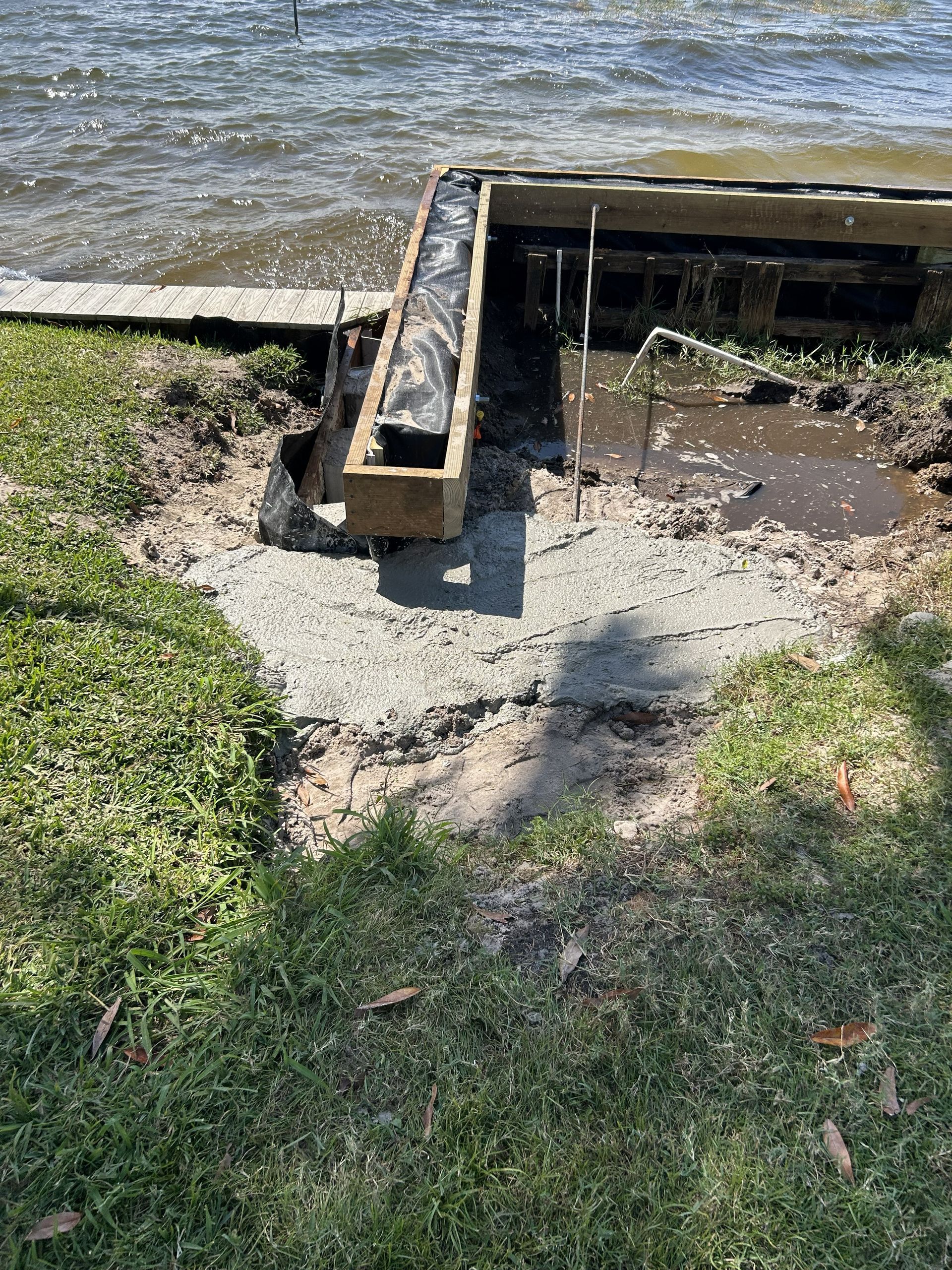 Damaged concrete dock with muddy water at the edge of a lake, beside grass and shoreline.