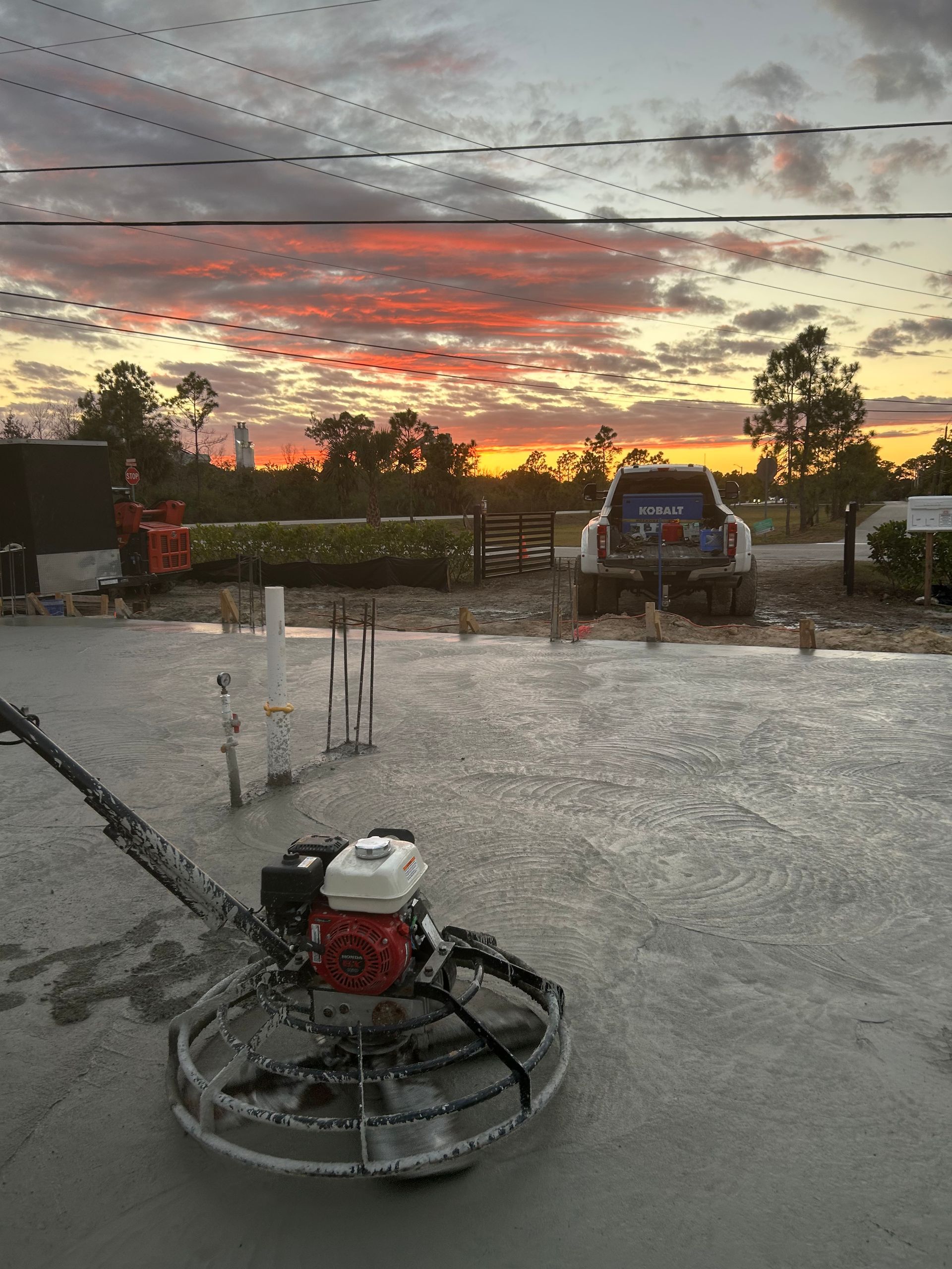 Concrete floor with a power trowel at sunset, water spraying under a colorful sky.