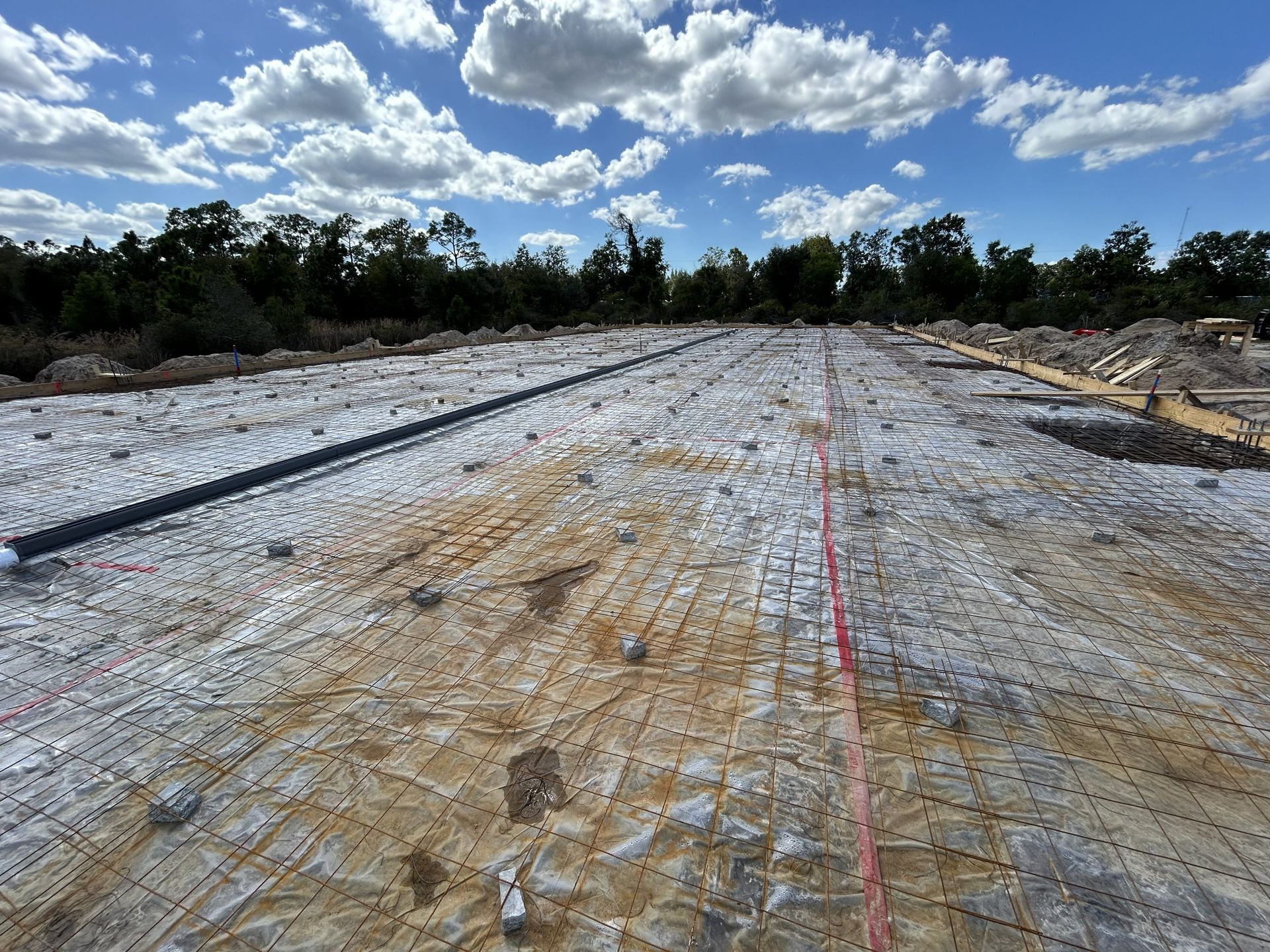 Gravel roadbed under construction with red survey line, bordered by trees beneath a cloudy blue sky