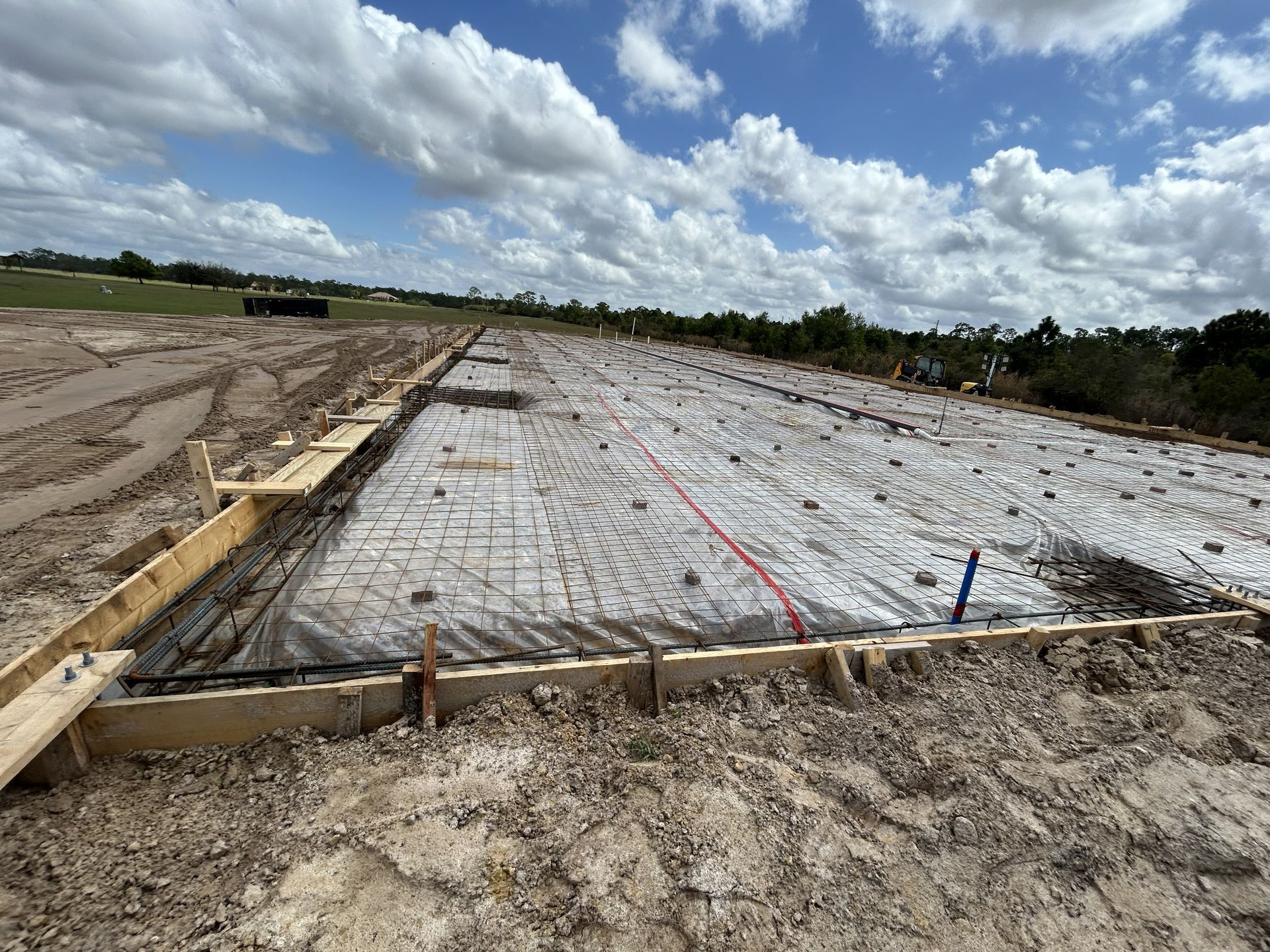 Concrete foundation slab under construction beside a dirt site, with wooden forms and rebar under a cloudy sky