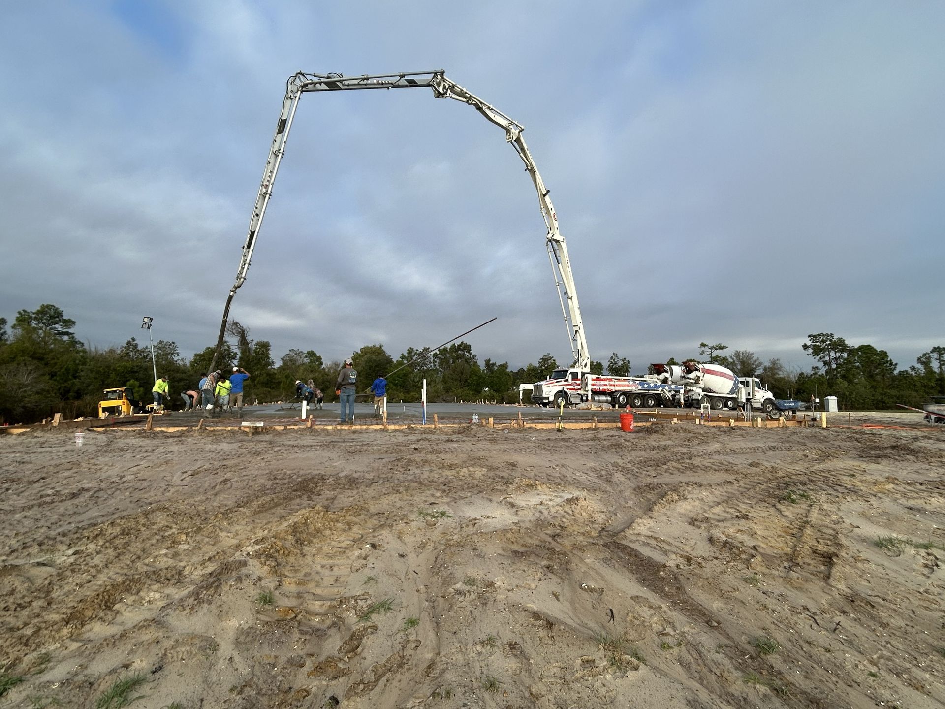 Concrete pump boom arcing over a muddy construction site with workers and trucks in the background