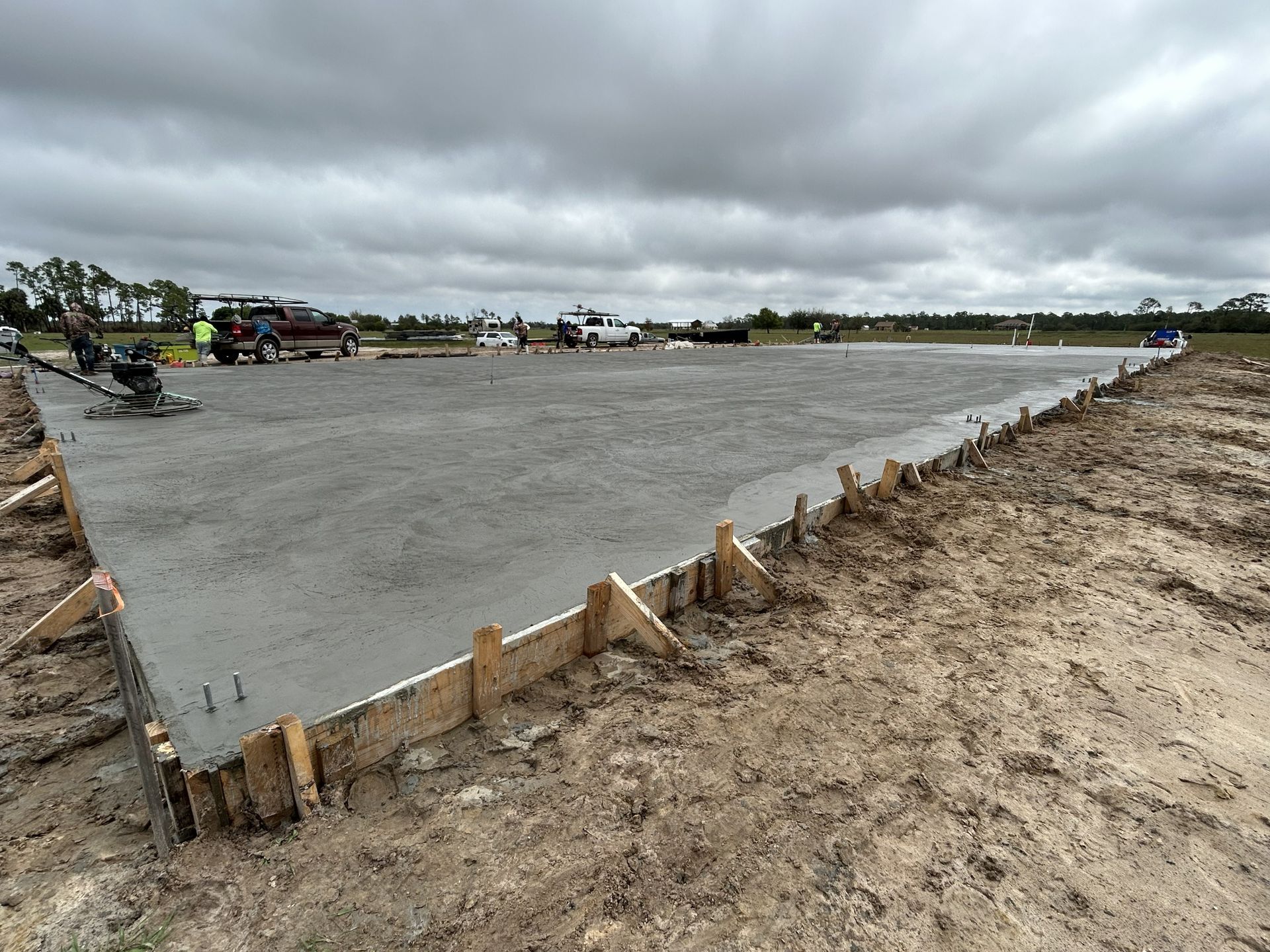 Freshly poured concrete slab at a construction site under a cloudy sky, bordered by wooden forms and dirt.