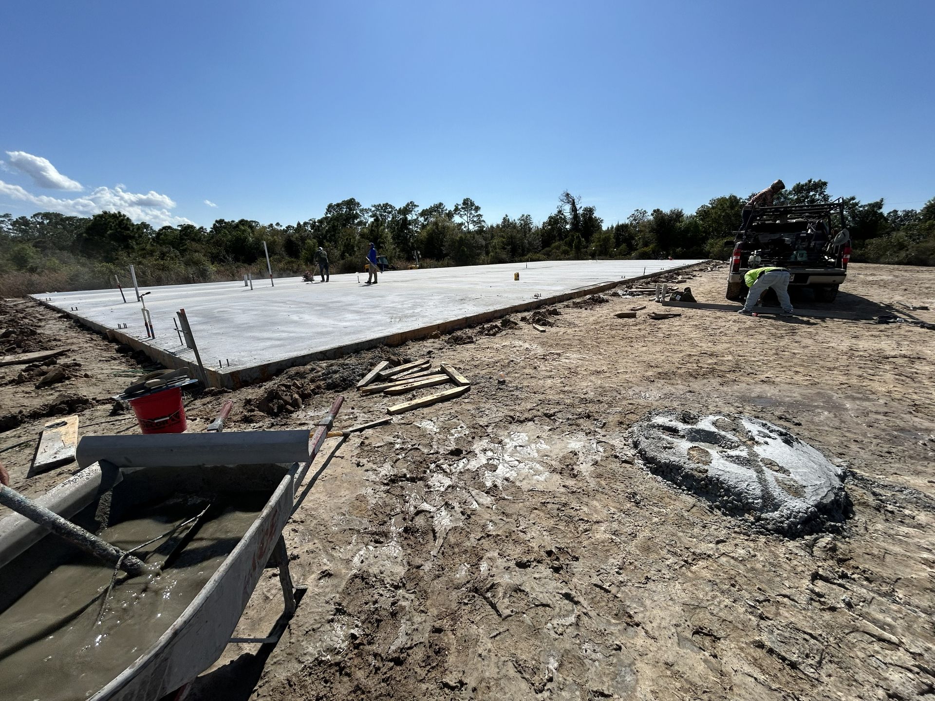 Workers pour concrete for a large slab on a sunny construction site, with tools and an ATV nearby.