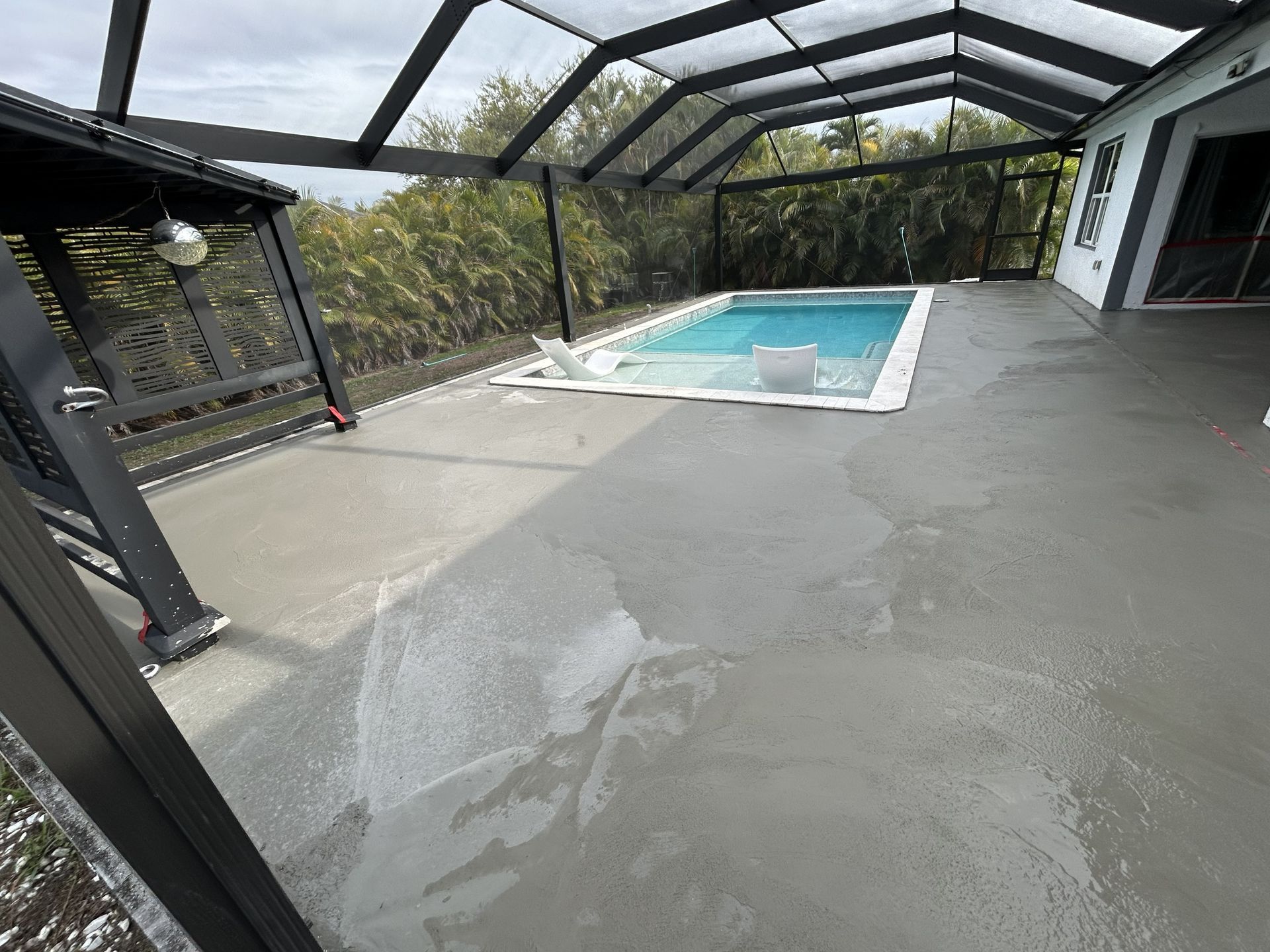 Covered backyard patio with a small swimming pool and wet concrete floor under a screened roof