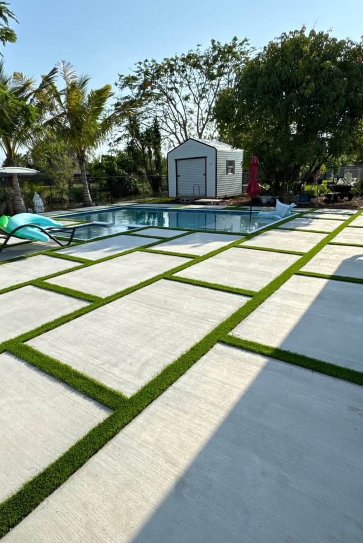 Modern backyard with a pool, white patio slabs, green grass strips, and a small shed under trees