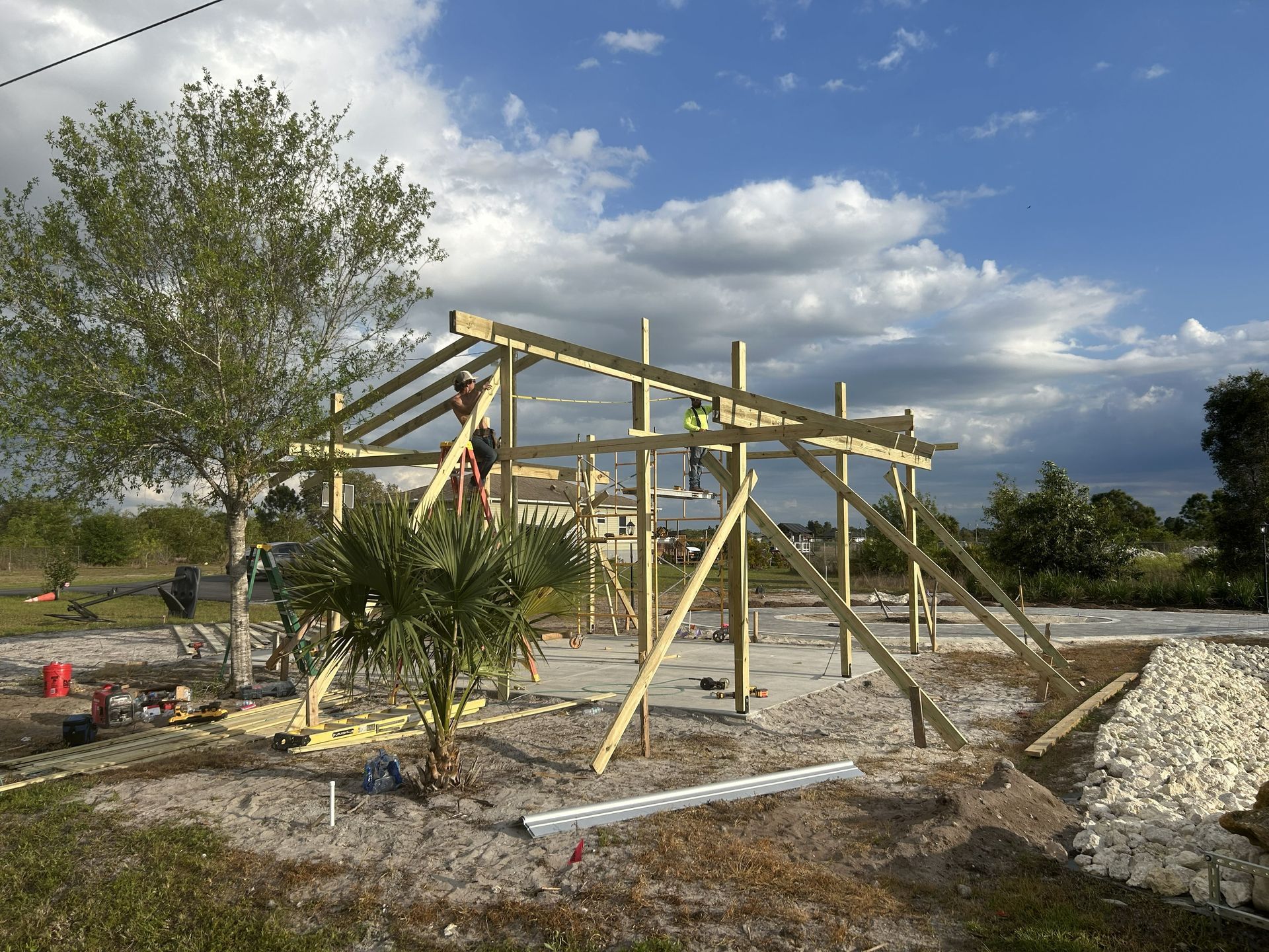 Wooden frame structure under construction on a gravel lot with cloudy sky and trees nearby