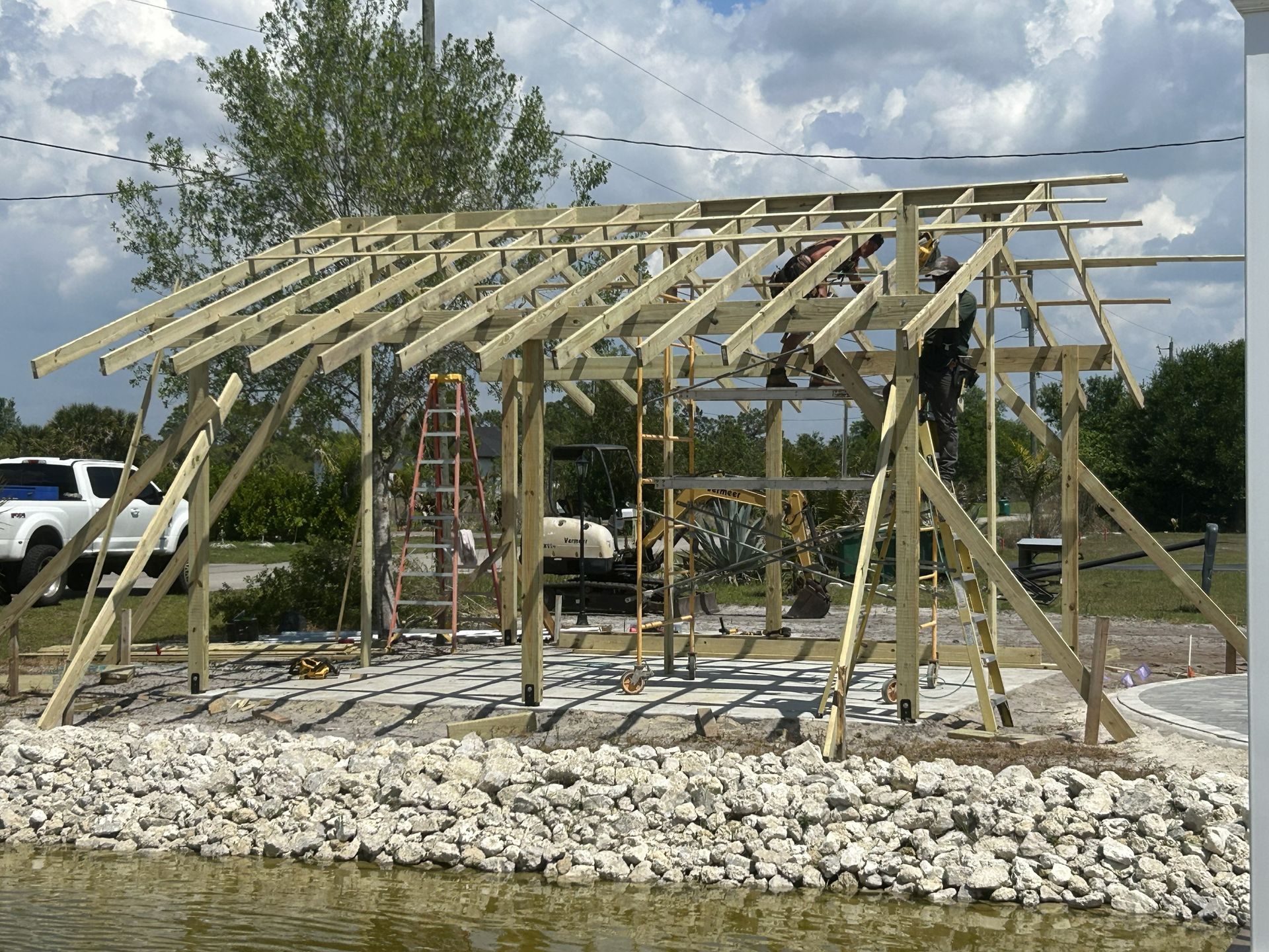 Wooden pavilion under construction on a rocky lot with a partly framed roof and cloudy sky.