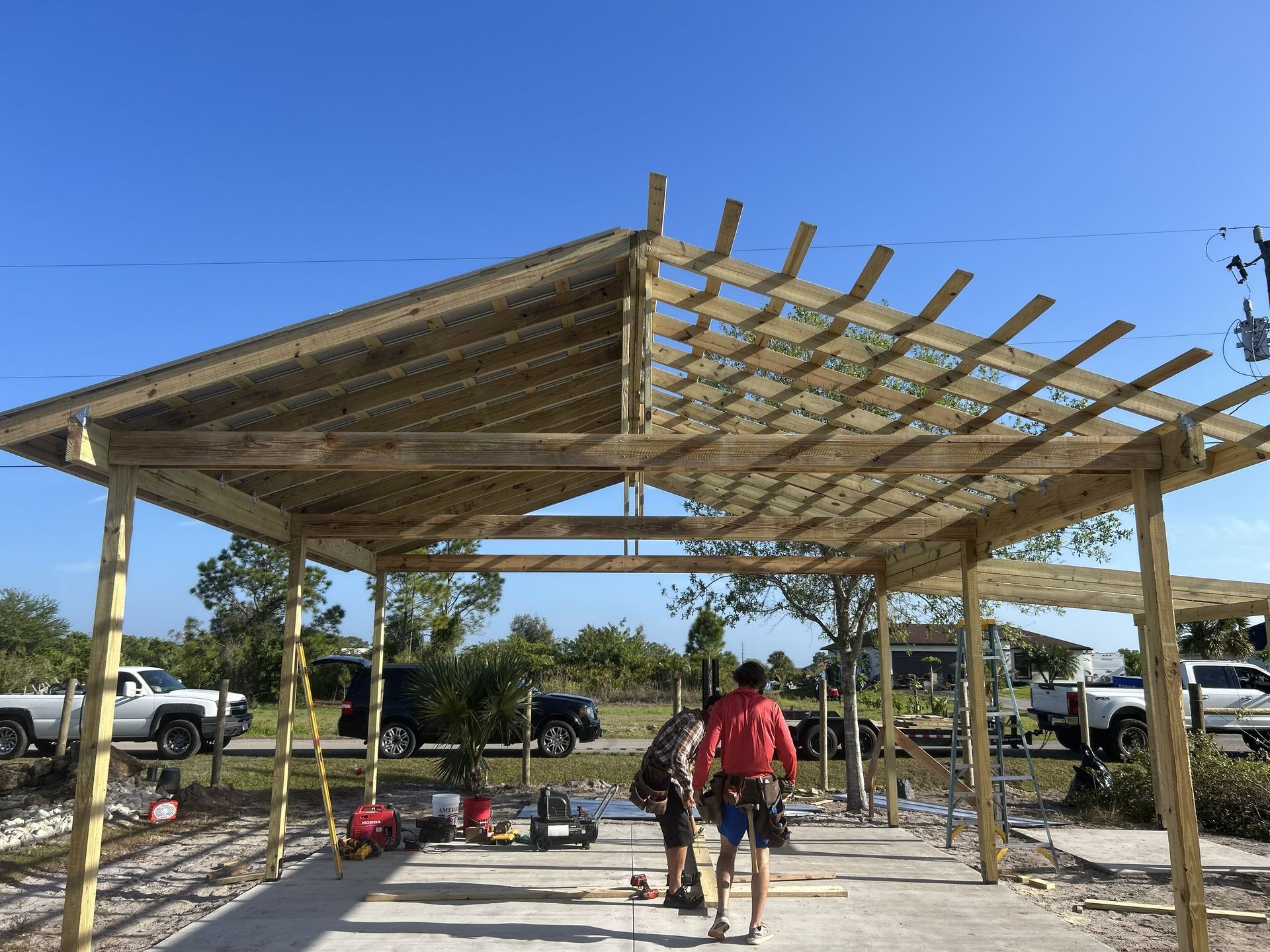 Partially built wooden pavilion with workers on a concrete slab under a clear blue sky