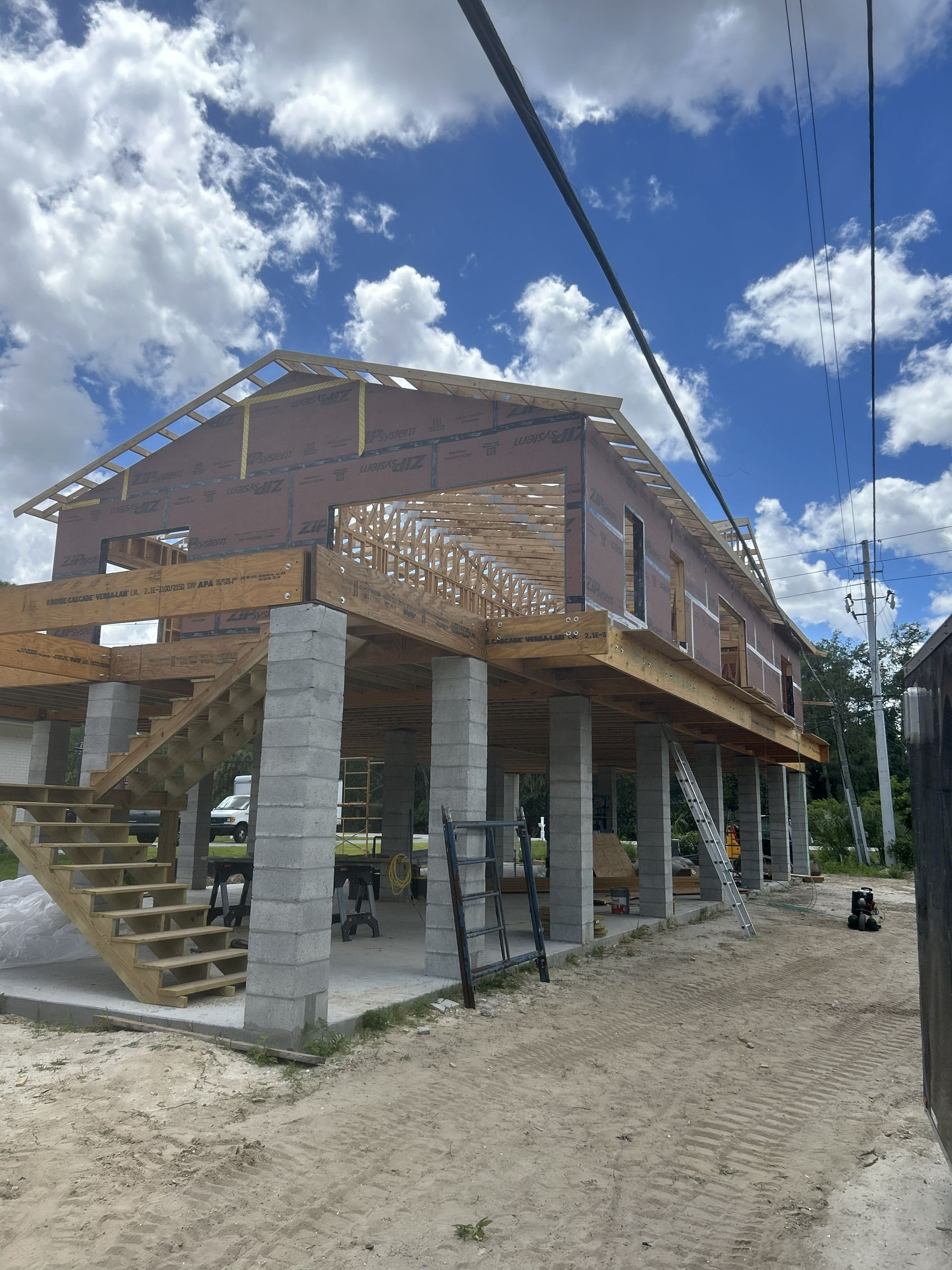 Elevated wooden house under construction on concrete pillars, with stairs and a gravel road beneath, under a blue sky