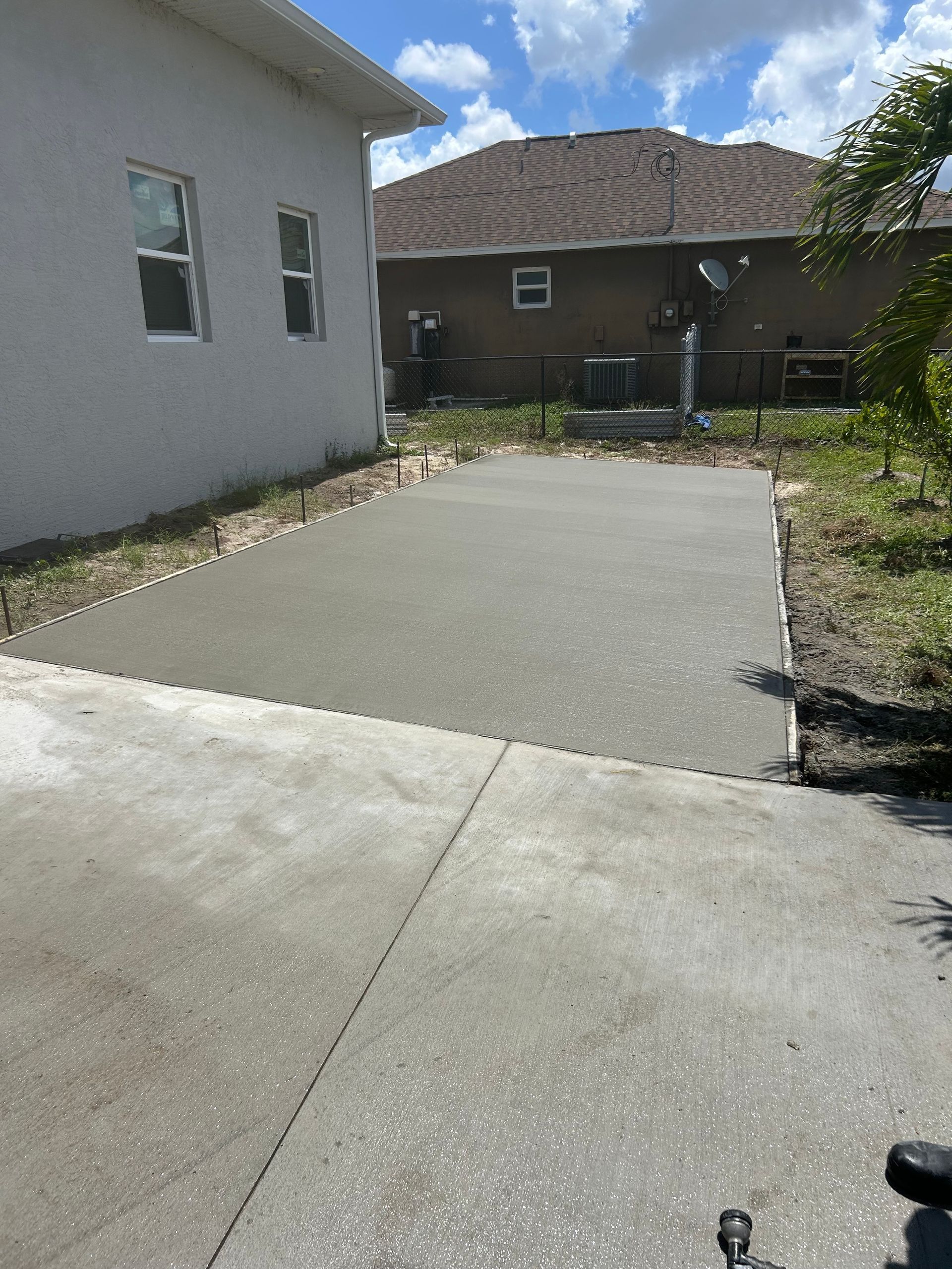 Concrete driveway and patio area beside a house, leading to a fenced backyard under a partly cloudy sky