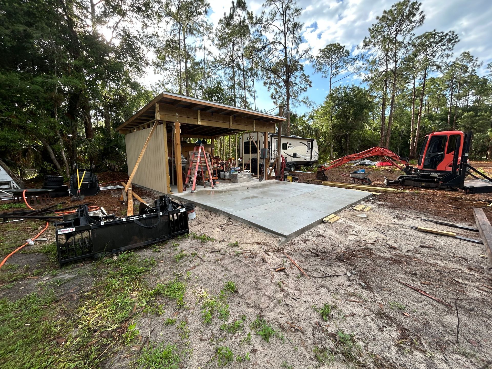 Construction site with concrete slab, shed, excavator, and muddy ground surrounded by trees