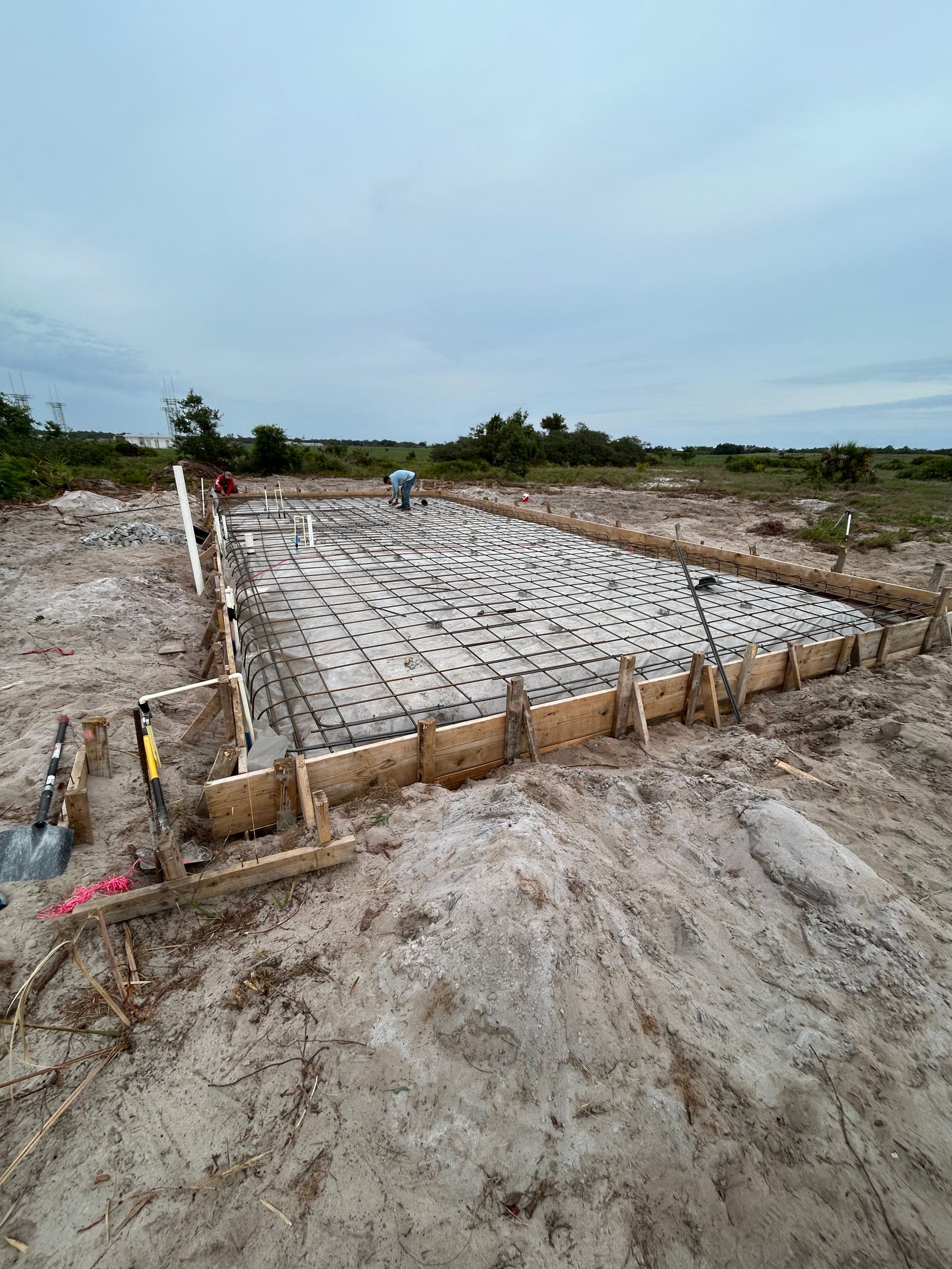 Concrete foundation slab under construction on a barren site, with wooden formwork and workers in the distance