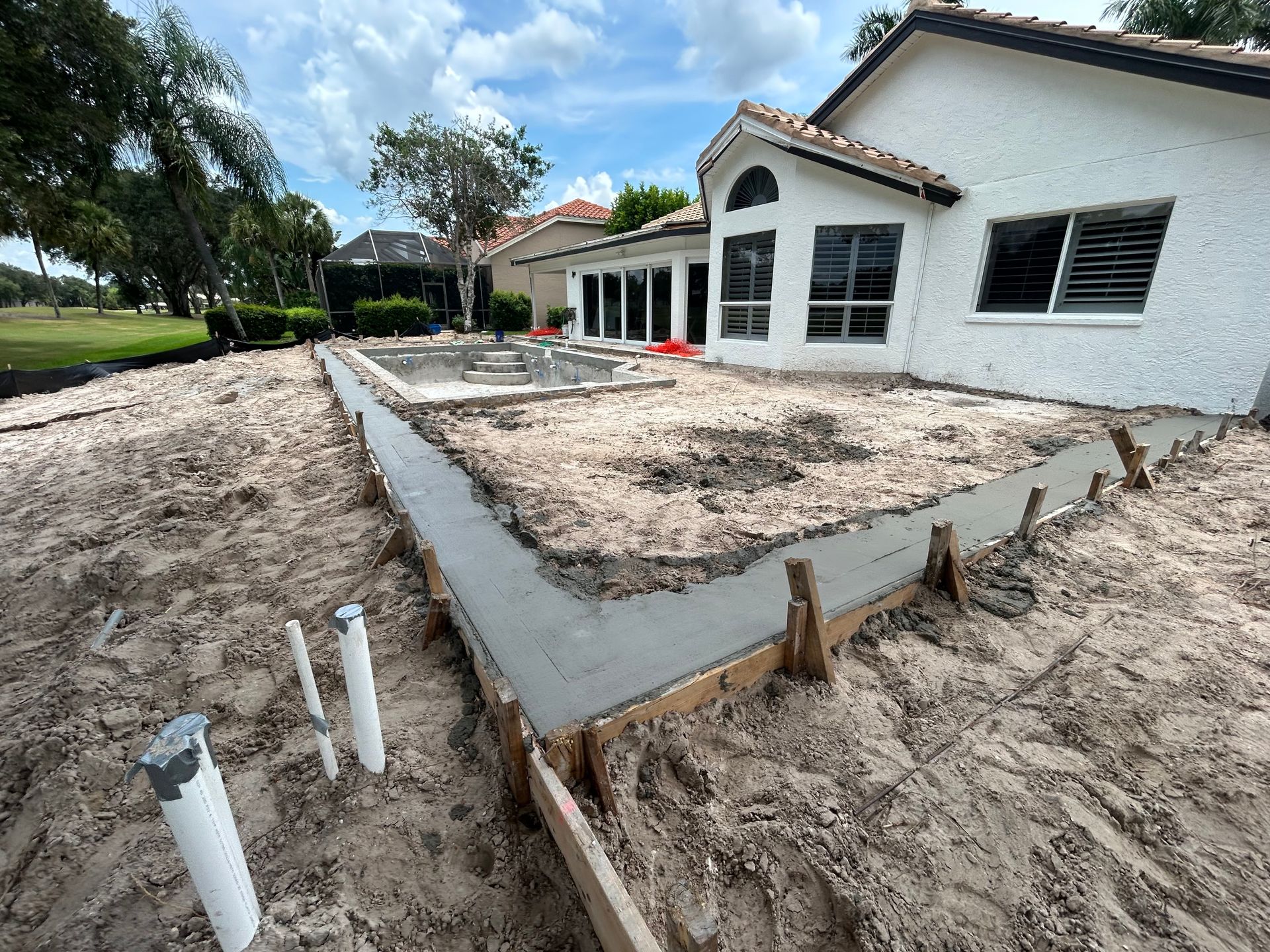 Fresh concrete foundation beside a white house, with sand, pipes, and landscaping work in progress