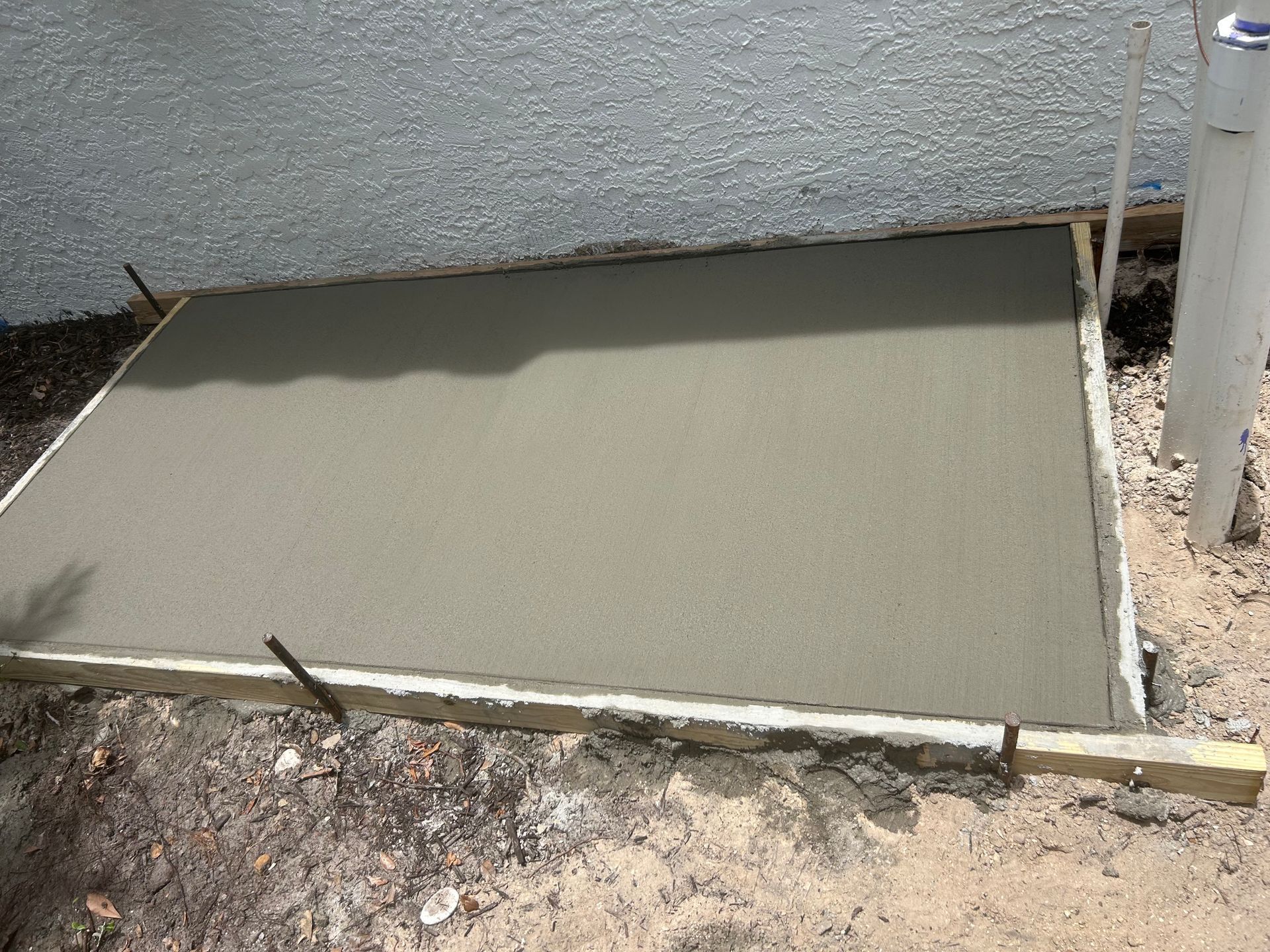 Freshly poured concrete slab beside a stucco wall, bordered by dirt and a white post.