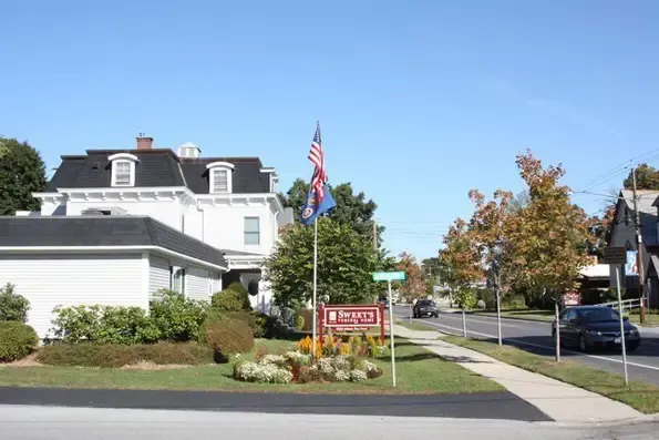 Street view with white building, sign, flags, and cars on road.
