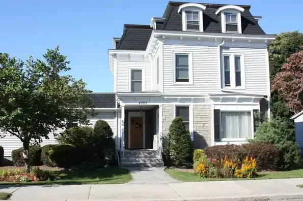 Three-story light gray house with a black roof, surrounded by trees and bushes on a sunny day.