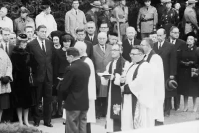 Mourners at a funeral. Priests in robes stand near a crowd of people in dark attire outdoors.