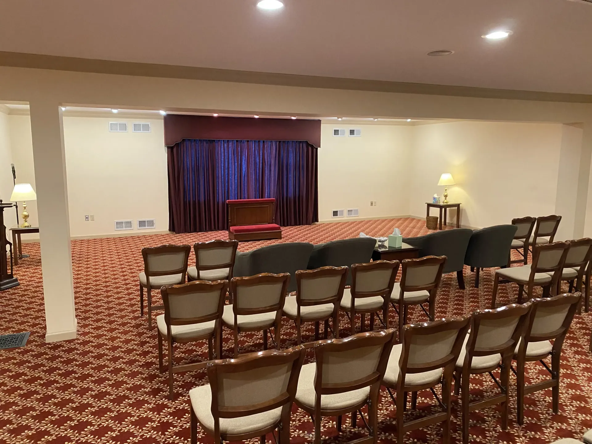 A beige room with rows of chairs facing a stage with maroon curtains. Red patterned carpet.